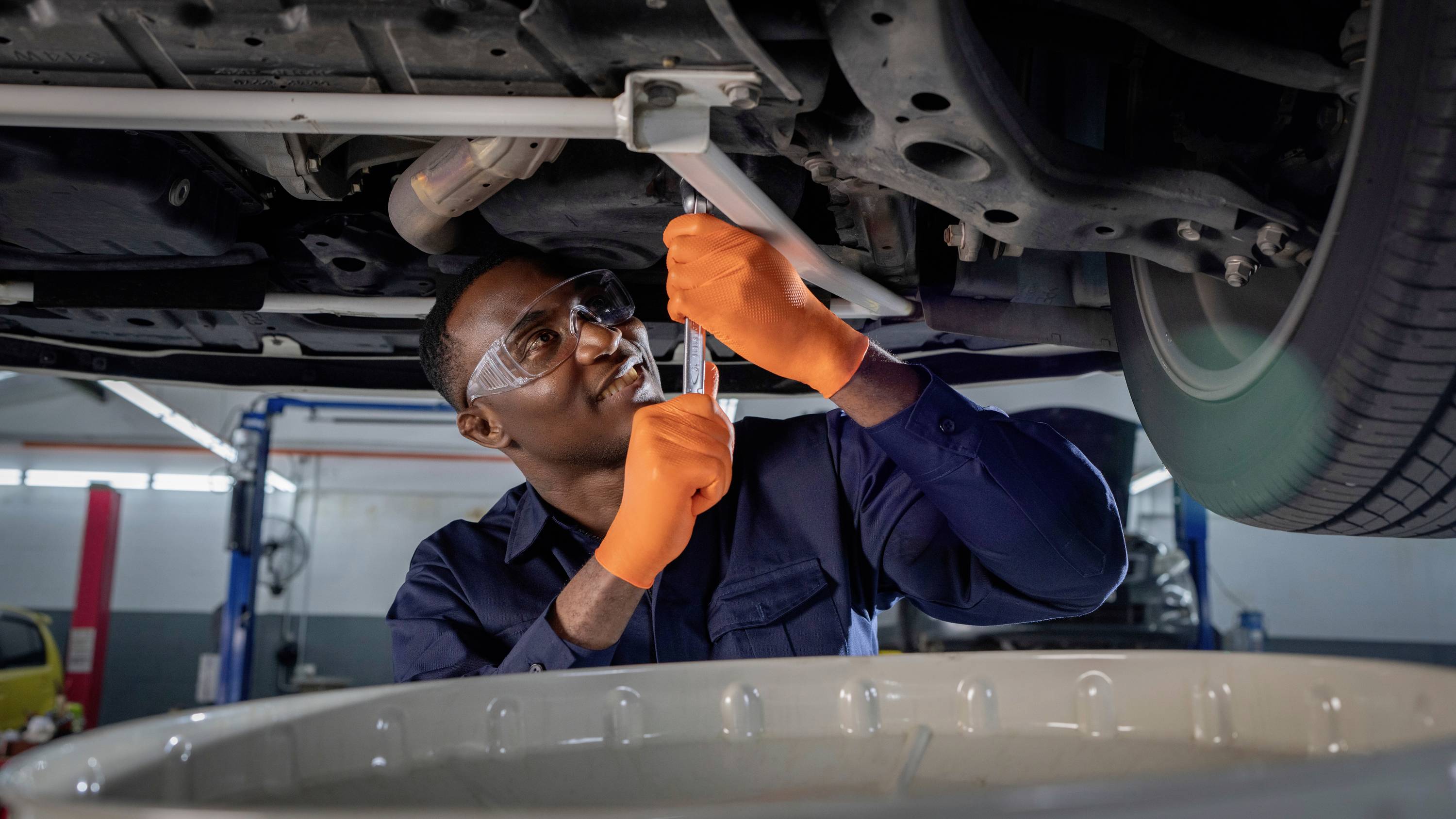 Ein Mechaniker mit Schutzbrille und orangefarbenen Handschuhen repariert ein Fahrzeug von unten in einer Werkstatt.