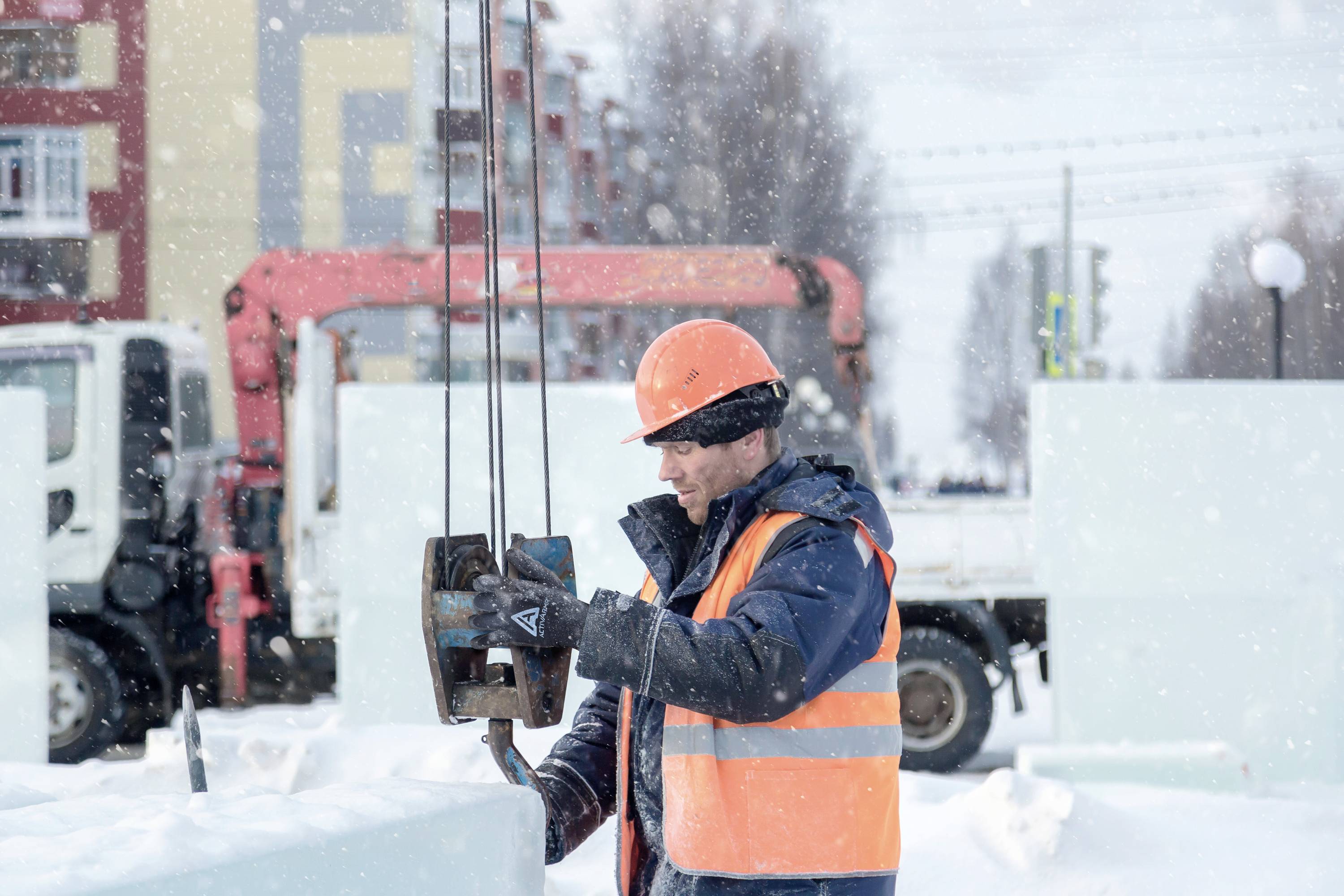 Ein Bauarbeiter mit Helm und Warnweste bedient Hebeausrüstung auf einer verschneiten Baustelle, umgeben von Gebäuden und LKWs.