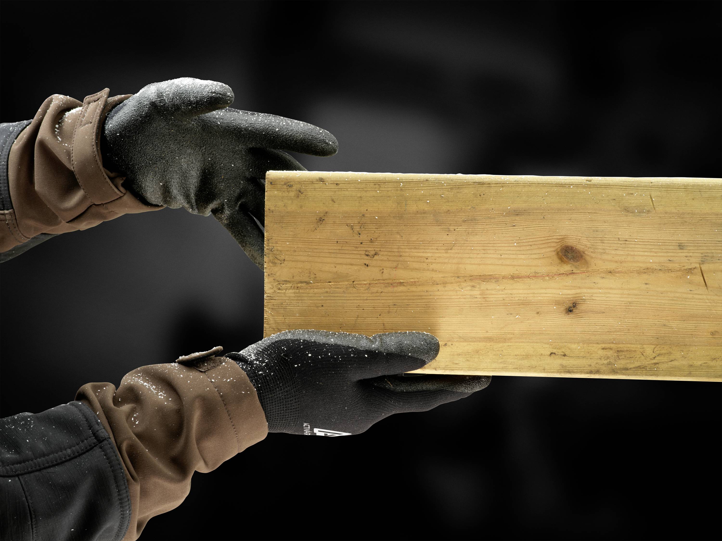 Zwei behandschuhte Hände halten ein Holzbrett fest. Der Hintergrund ist unscharf und dunkel, was den Fokus auf das Brett legt.