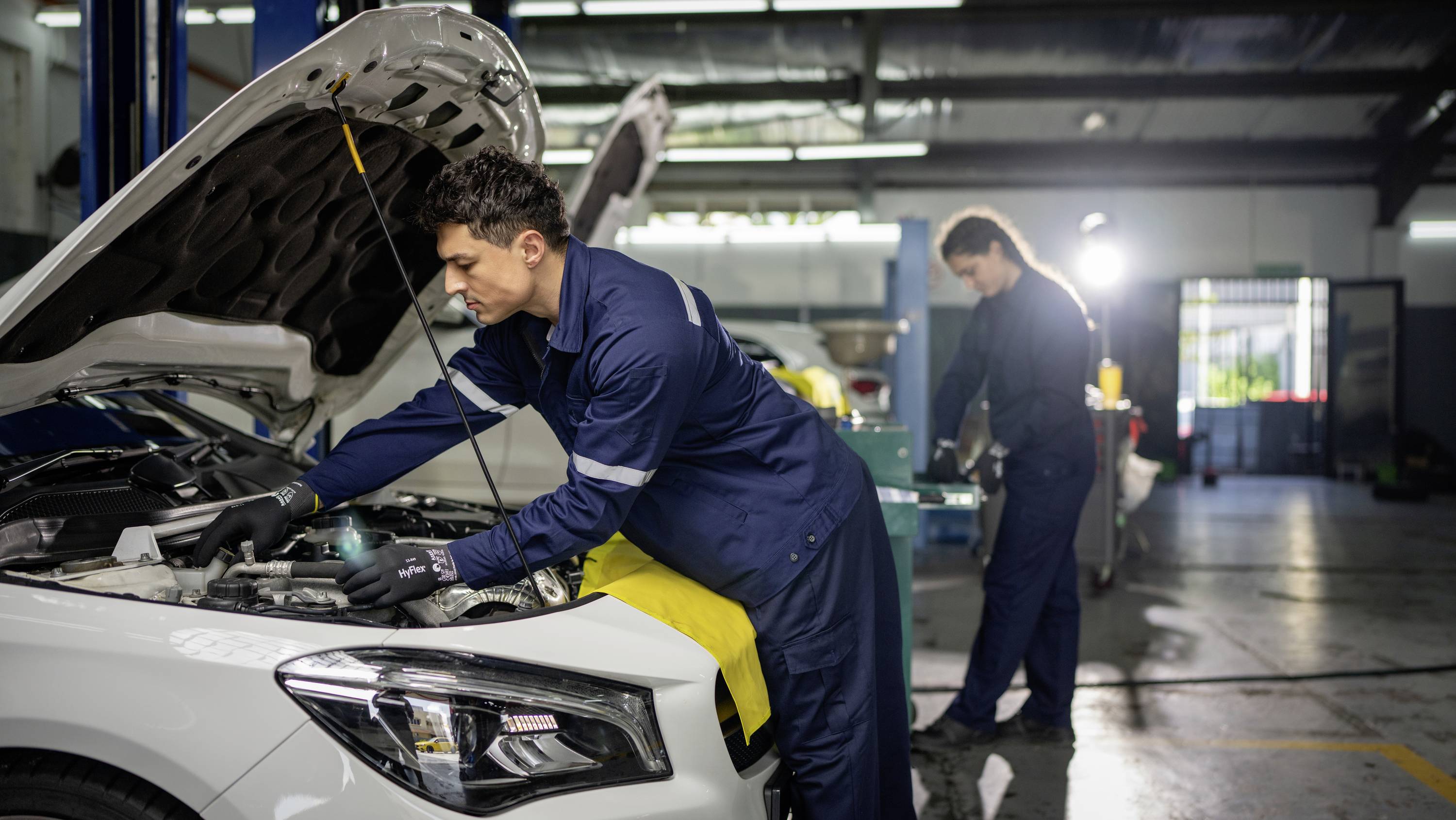 Ein Mechaniker arbeitet unter der Motorhaube eines Autos in einer Werkstatt, während ein zweiter Mechaniker im Hintergrund beschäftigt ist.