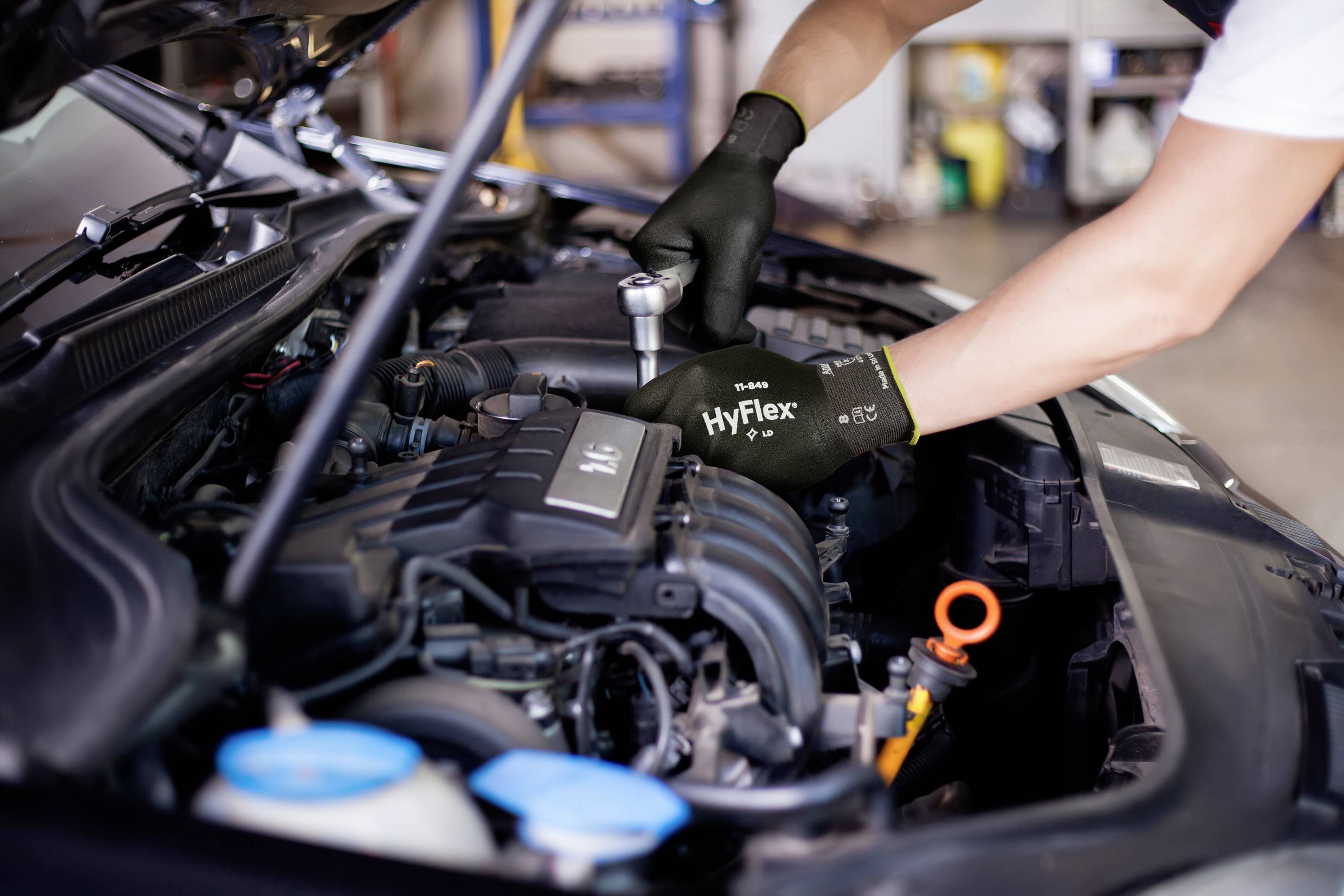 Eine Person in Handschuhen arbeitet am Motor eines Autos, das die Motorhaube geöffnet hat. Im Vordergrund befinden sich Werkzeuge und Motorflüssigkeiten.