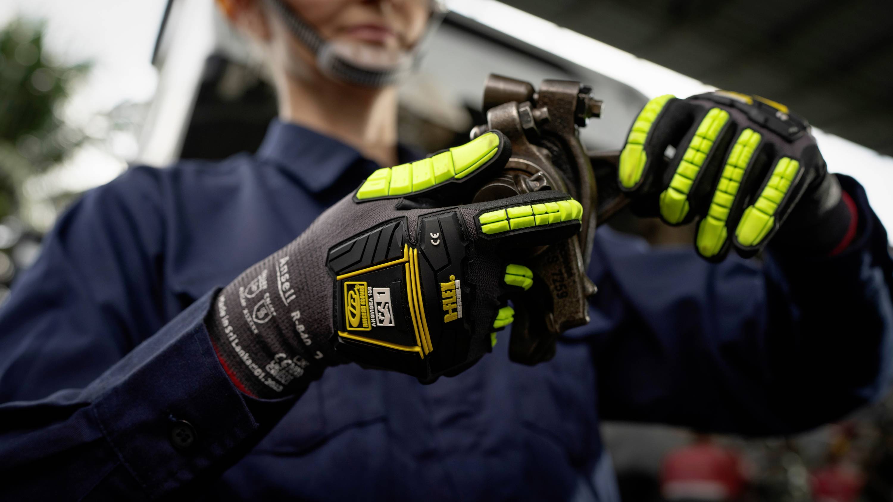Person in Arbeitskleidung mit Schutzhandschuhen hält ein mechanisches Teil. Fokus auf die Handschuhe, die neonfarbene Details haben.