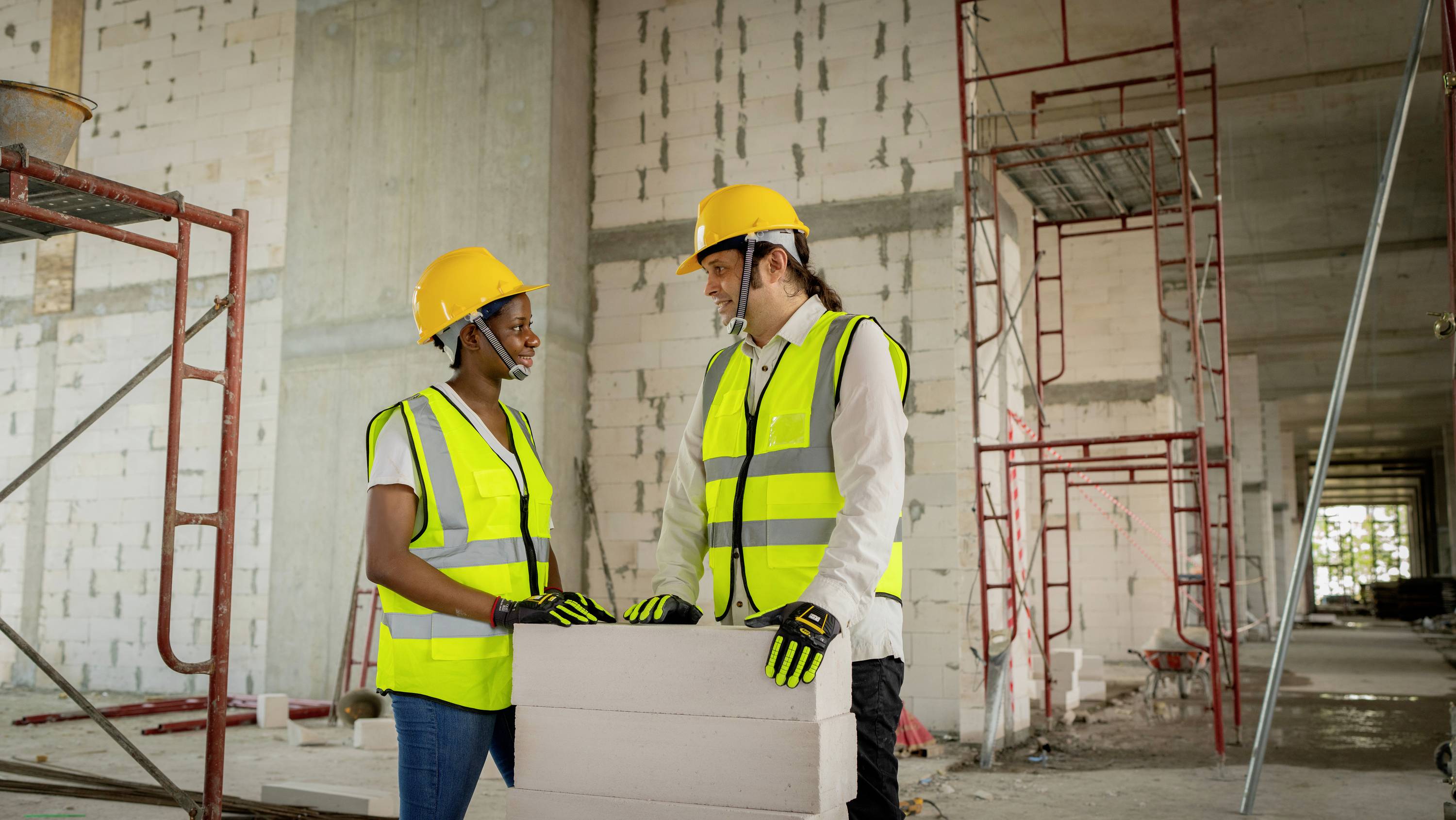 Zwei Bauarbeiter mit Schutzhelmen und Warnwesten sprechen in einem Rohbau. Sie halten Pläne in der Hand. Hintergrund zeigt Gerüste.