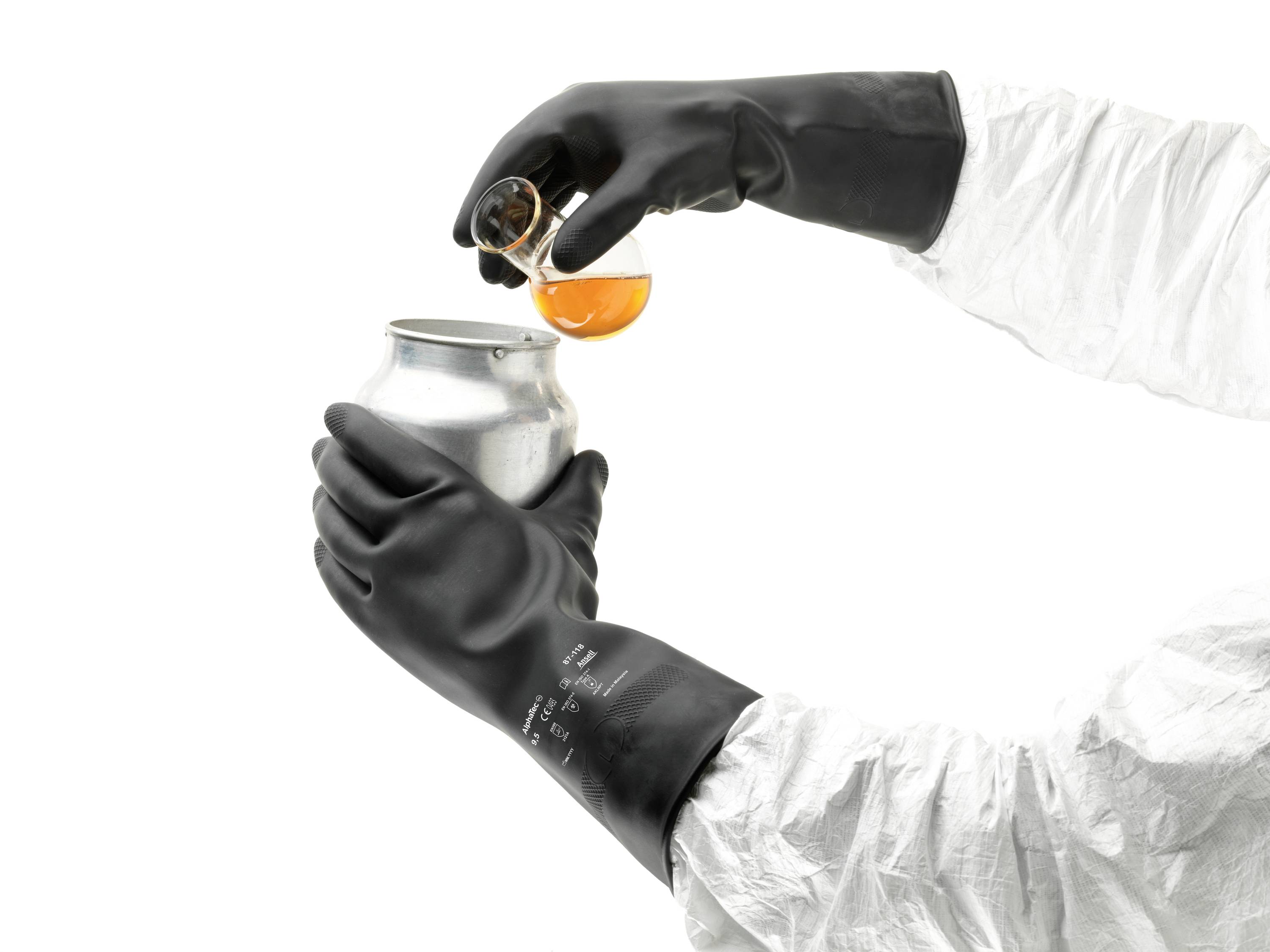 Eine Person in Schutzausrüstung gießt orange Flüssigkeit aus einem Becherglas in eine Metalldose. Hände tragen schwarze Handschuhe.