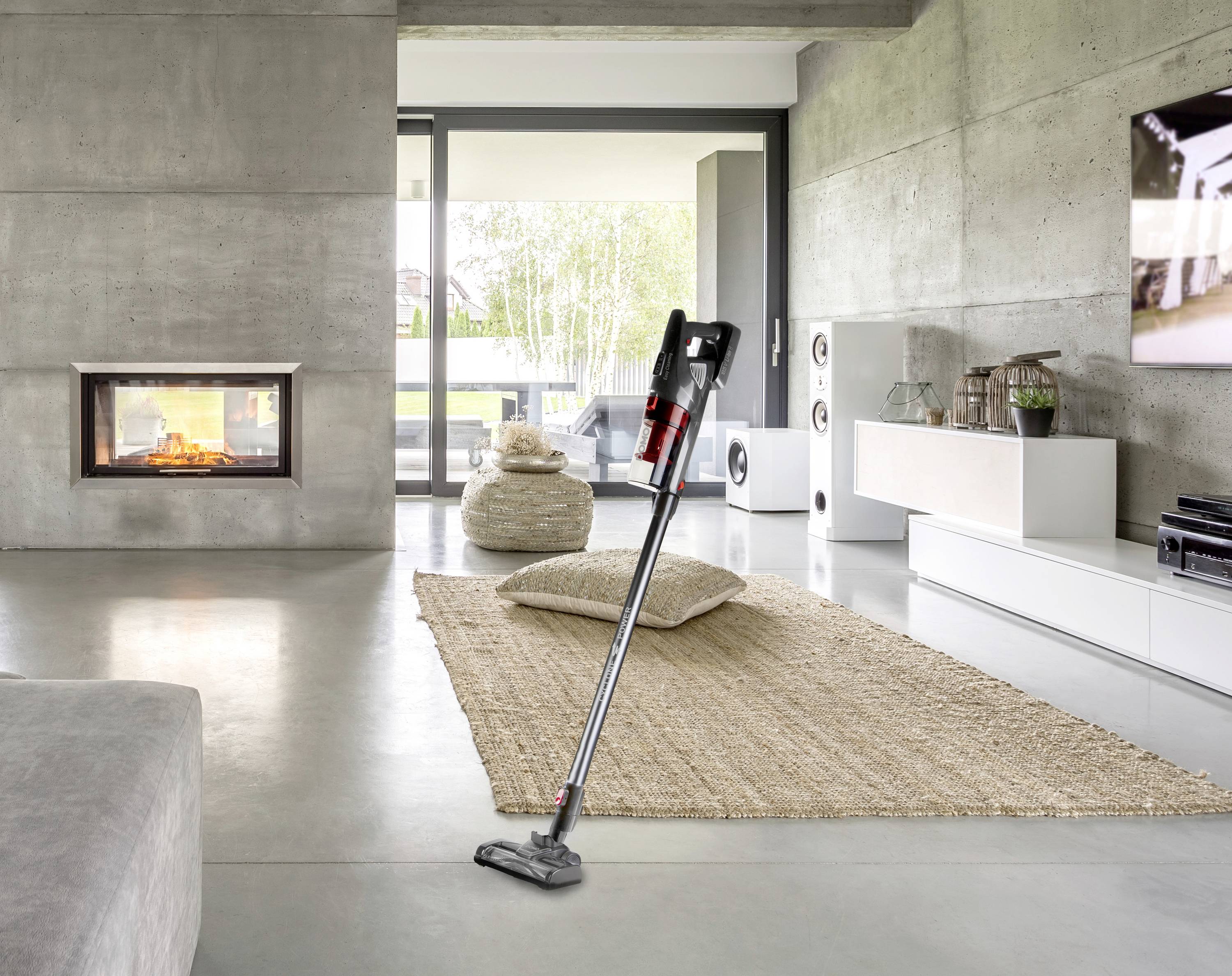 Ein kabelloser Staubsauger steht in einem modernen Wohnzimmer auf einem Teppich. Der Raum hat Betonwände und einen Kamin.