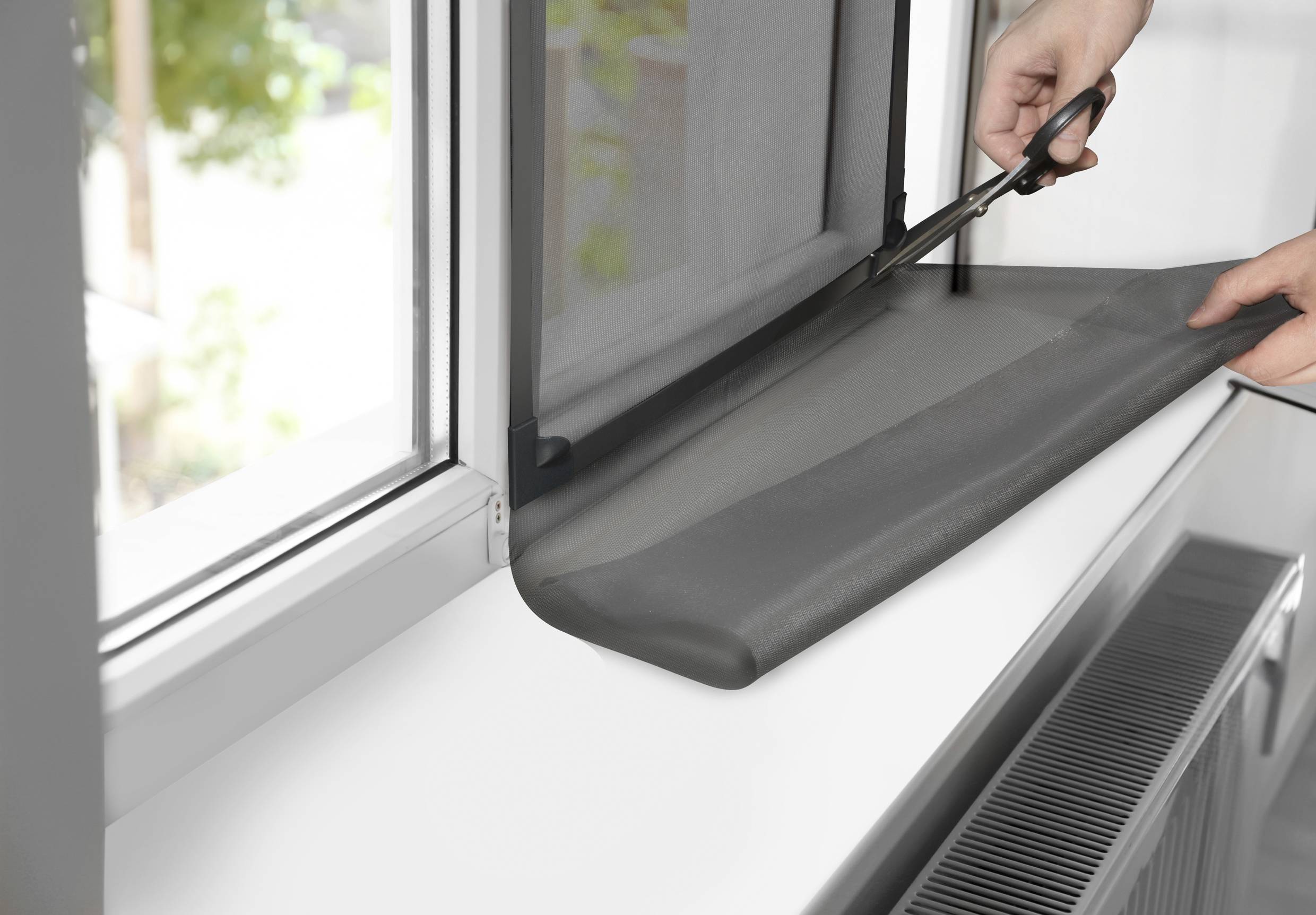 Eine Person schneidet mit einer Schere ein Fliegengitter für ein geöffnetes Fenster passend zu. Im Hintergrund sind verschwommen Bäume sichtbar.