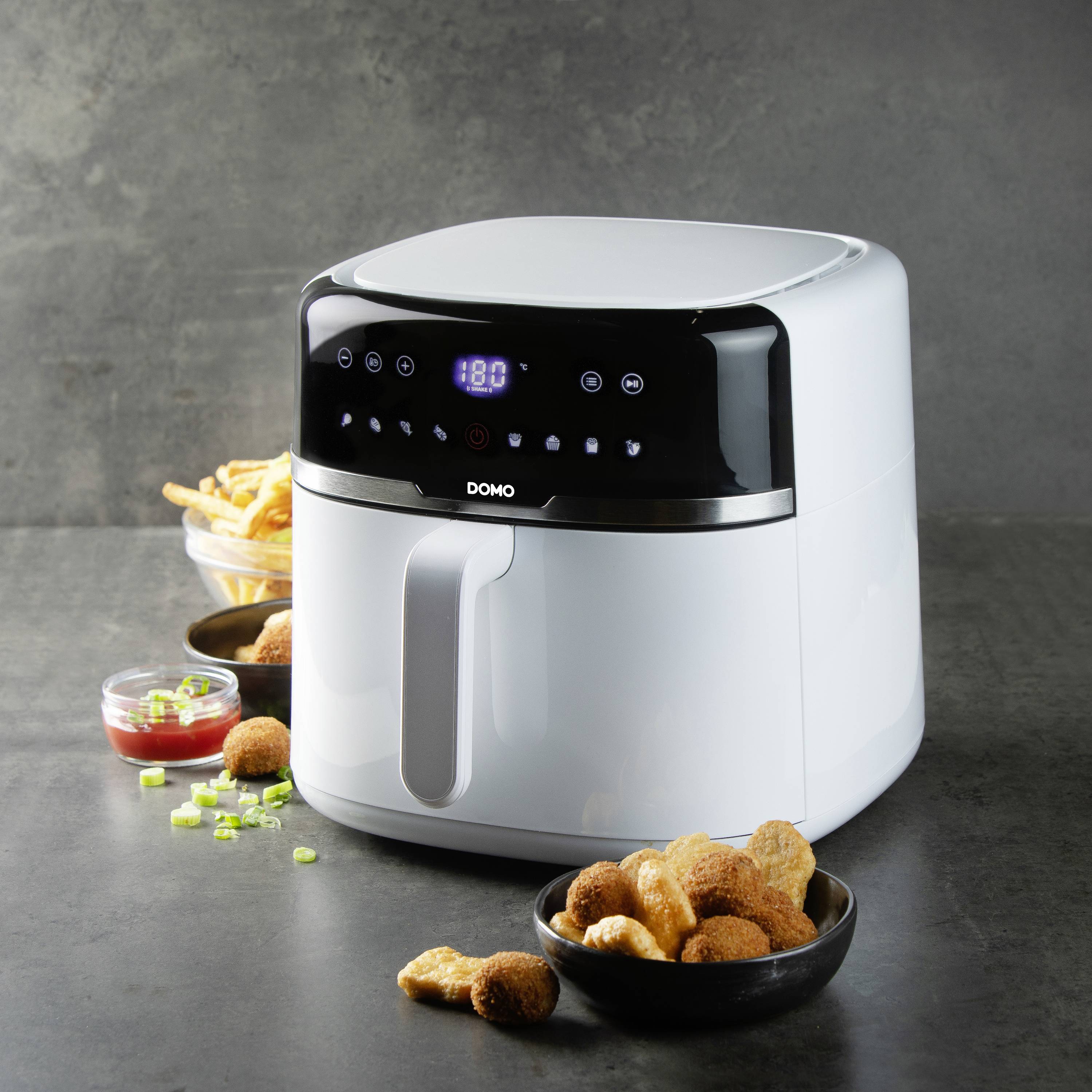Eine Heißluftfritteuse auf einem Tisch, umgeben von frittierten Snacks und Frühlingszwiebeln, zeigt eine Temperaturanzeige von 180 Grad.
