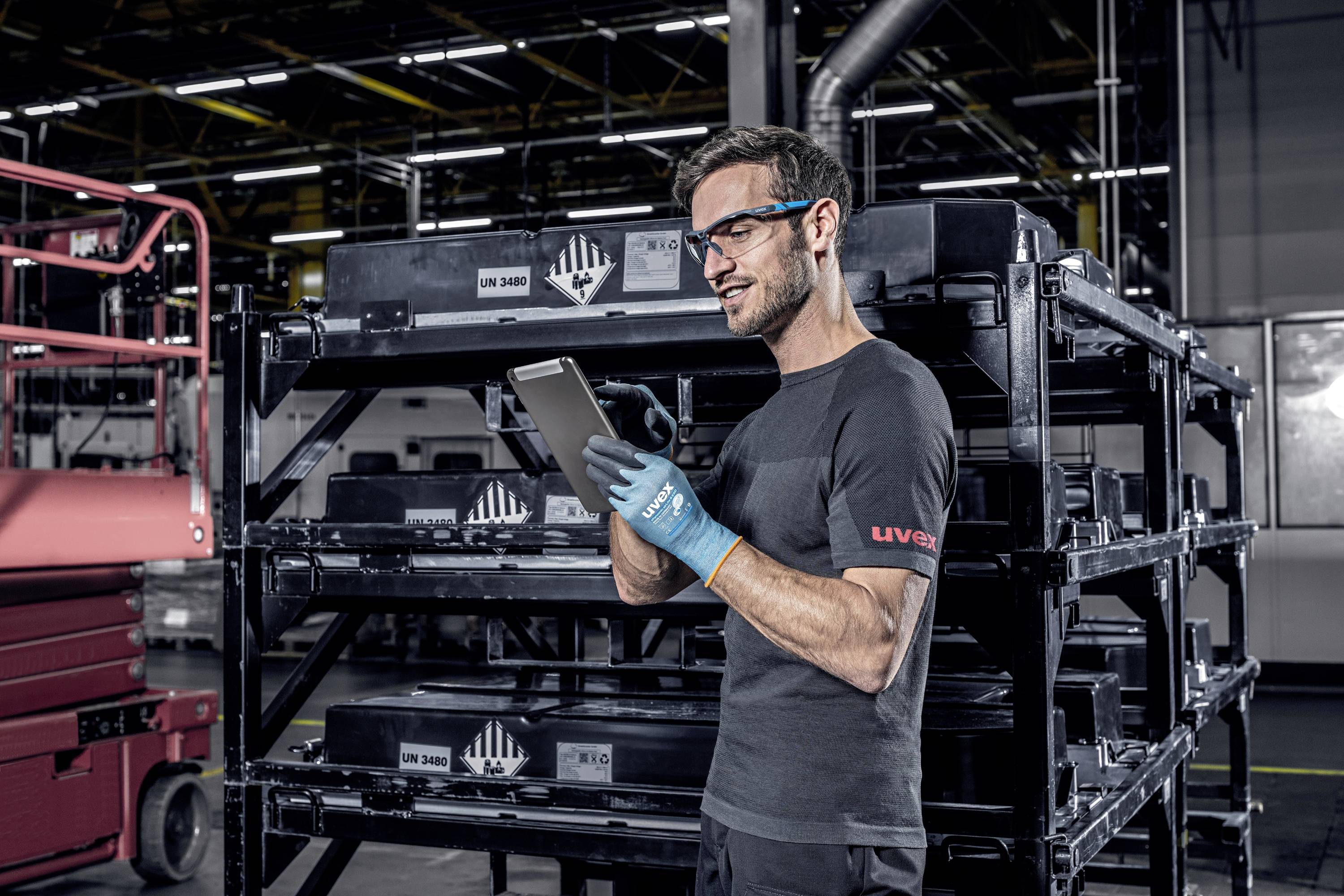 Ein Arbeiter in einem Lager trägt Schutzbrille und Handschuhe, benutzt ein Tablet und steht vor Regalen mit Kisten.