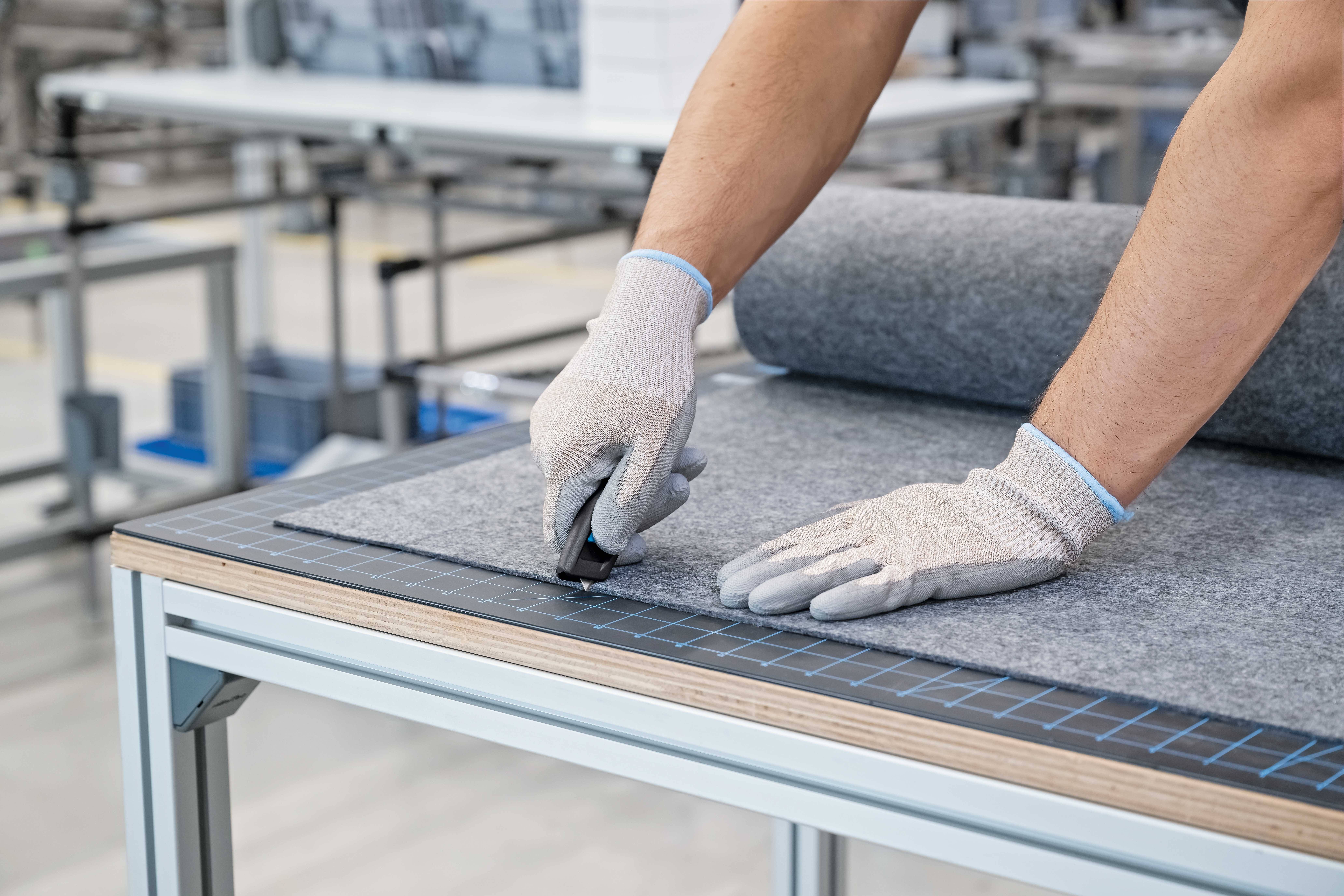 Eine Person schneidet mit einem Messer sorgfältig einen grauen Stoff auf einem Arbeitstisch. Sie trägt helle Schutzhandschuhe.