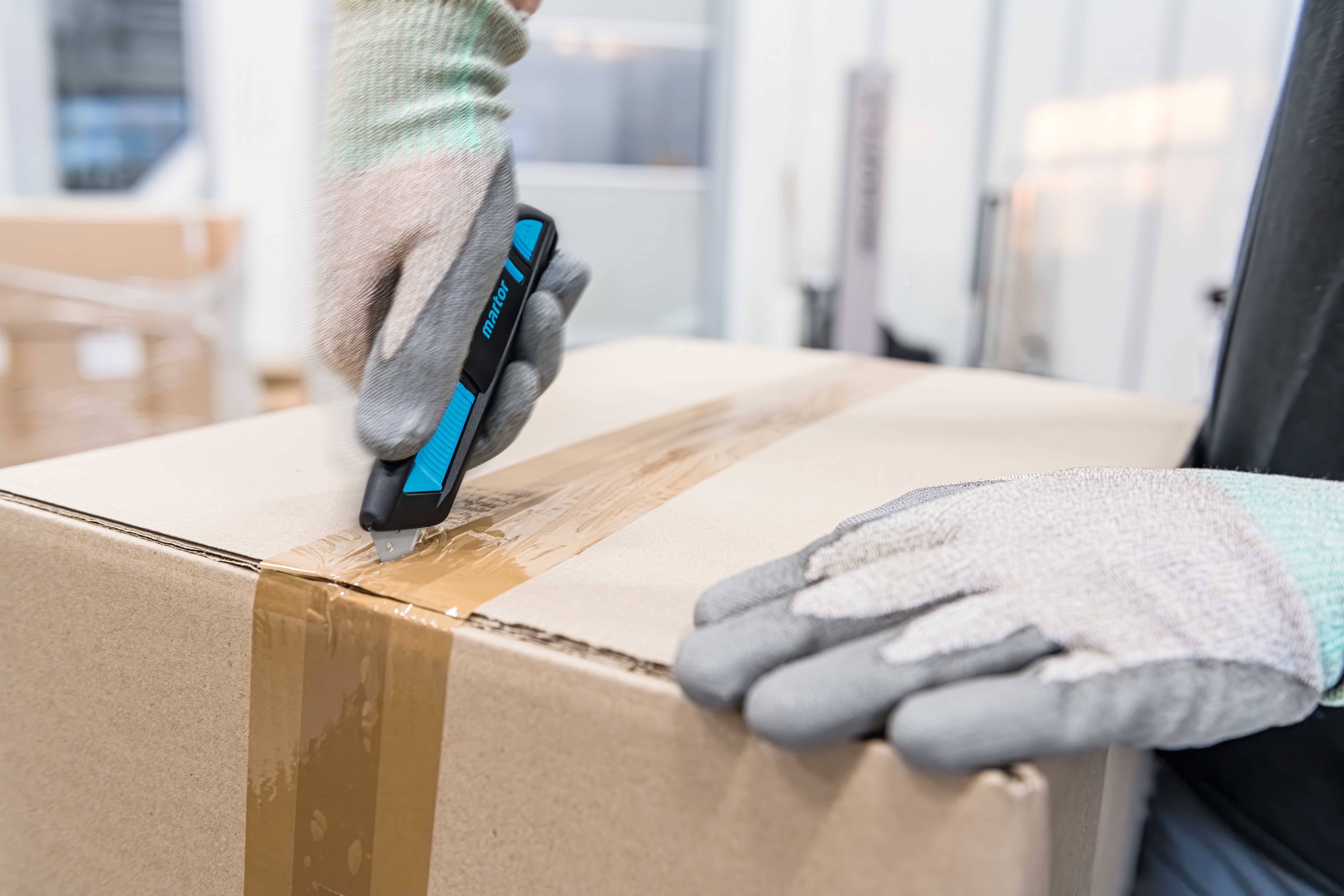 Eine Person öffnet mit einem Messer einen Karton, während sie Handschuhe trägt. Die Szene findet in einer Lagerumgebung statt.