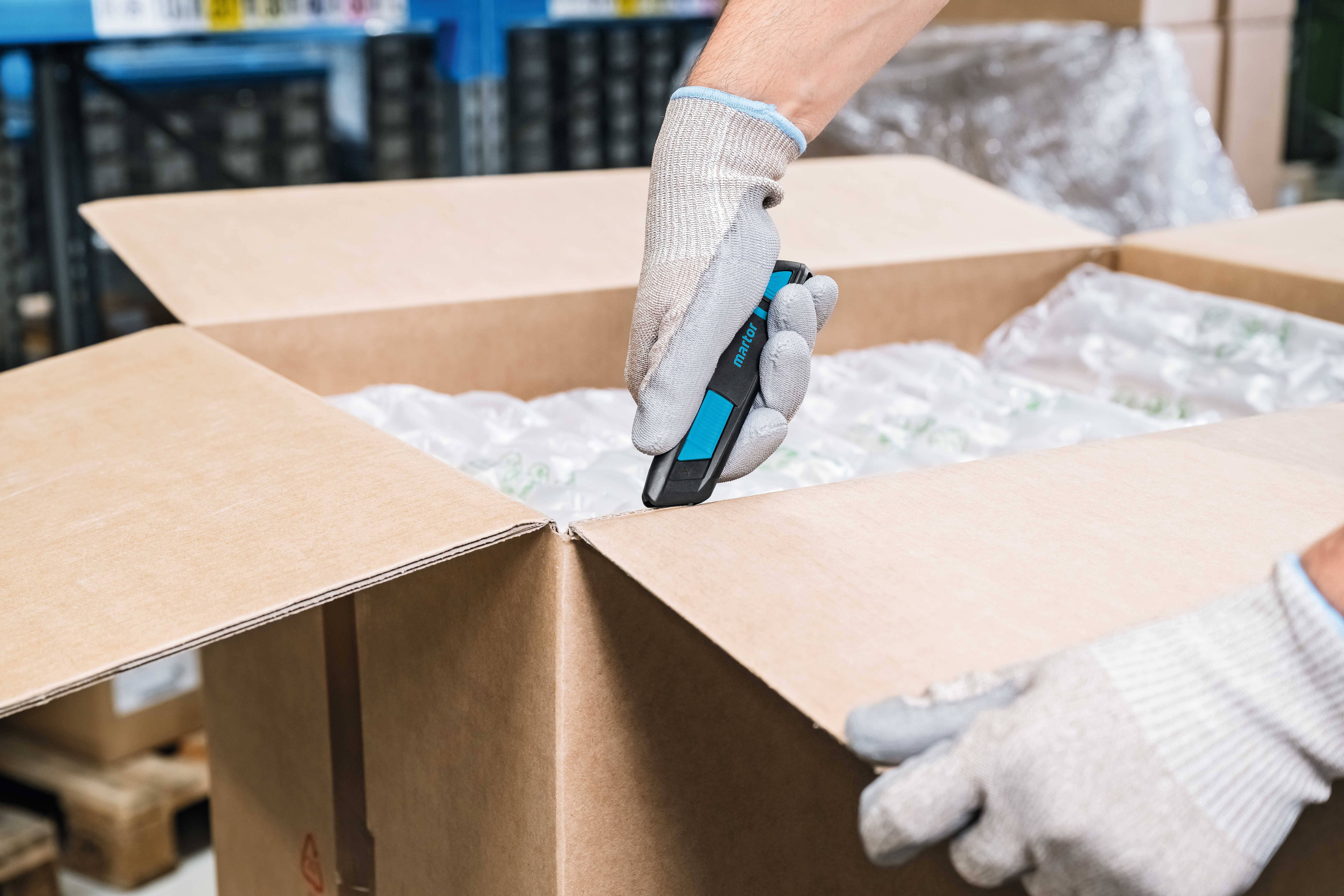 Eine Person öffnet mit einem Cutter in Handschuhen einen großen Karton, der mit Luftpolsterfolie gefüllt ist.