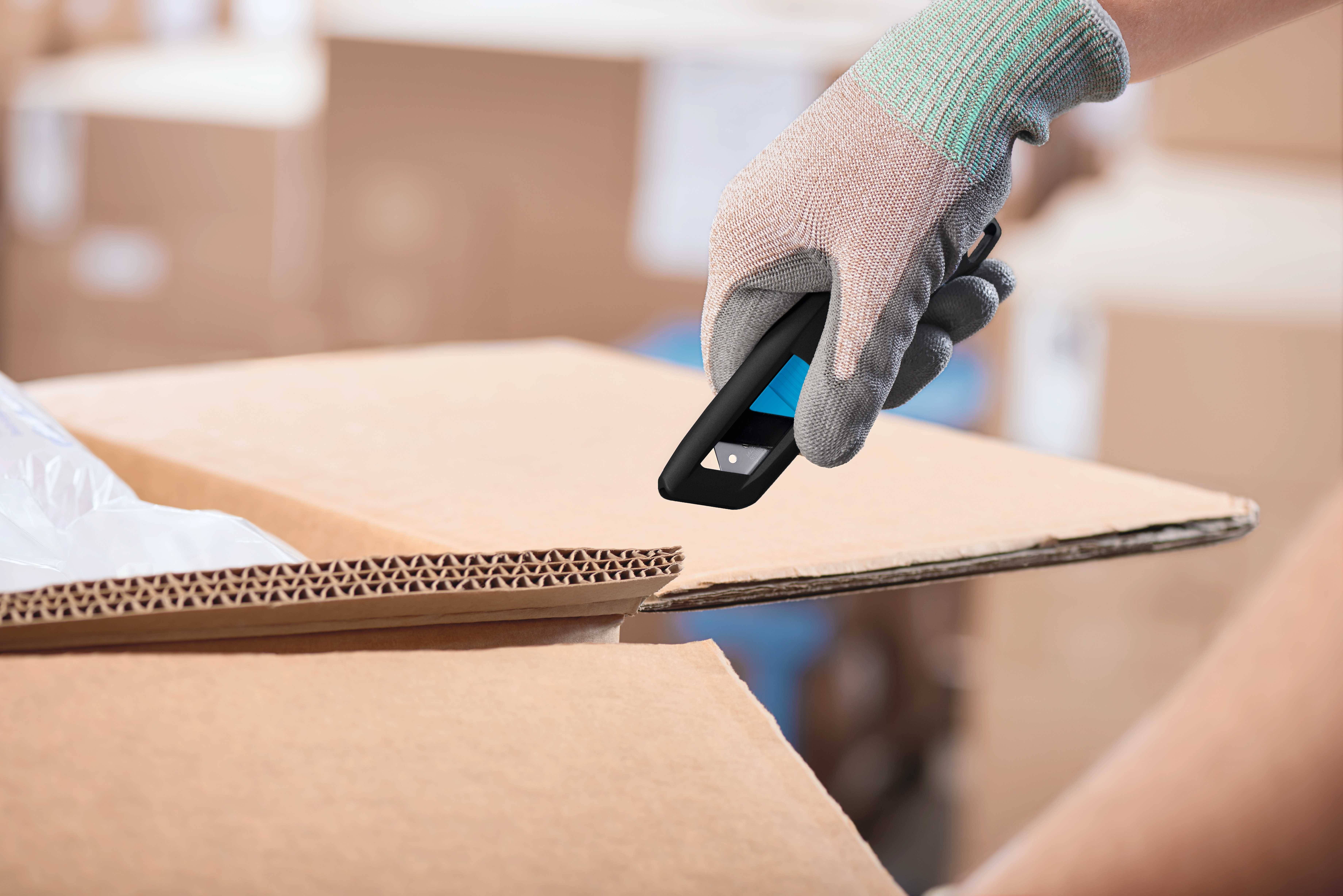 Hand mit Handschuh schneidet einen Karton mit einem Cutter-Messer, umgeben von mehr Kartons im Hintergrund.