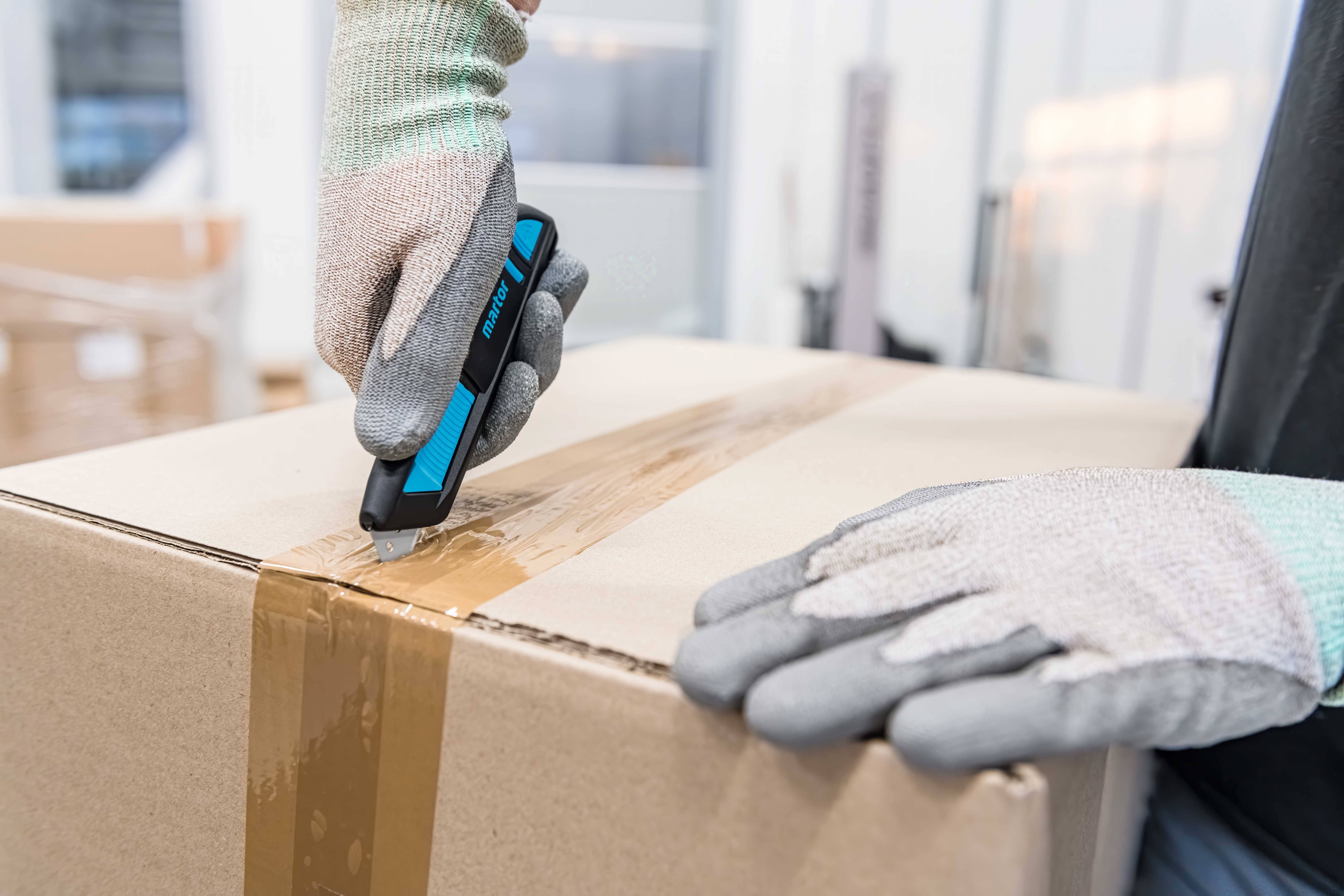 Person öffnet mit einem Messer ein braunes Paket, trägt graue Handschuhe, im Hintergrund unscharfe Lagerumgebung.