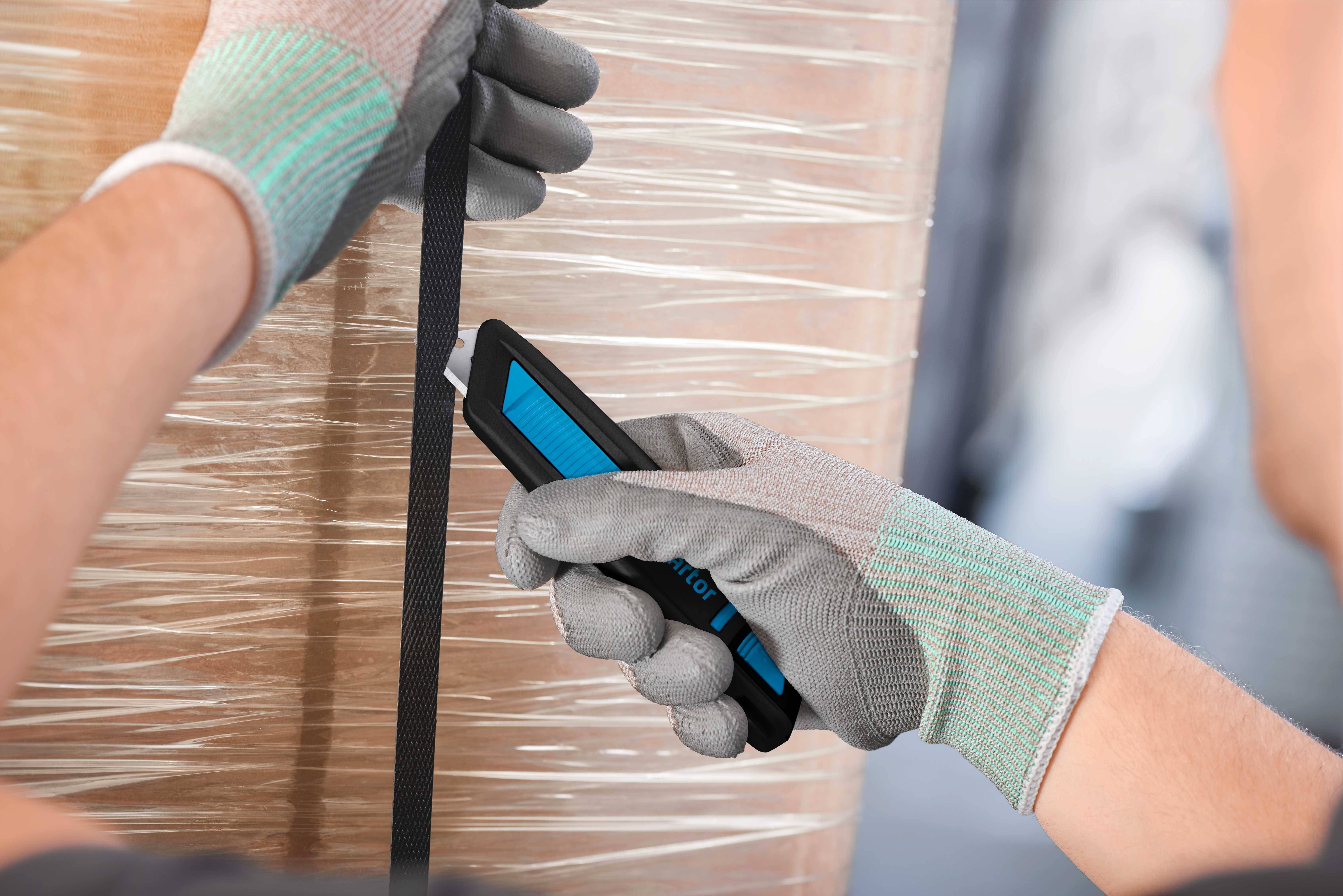 Person mit Handschuhen hält ein Cuttermesser und schneidet ein schwarzes Band, das um einen großen, in Folie verpackten Gegenstand gewickelt ist.