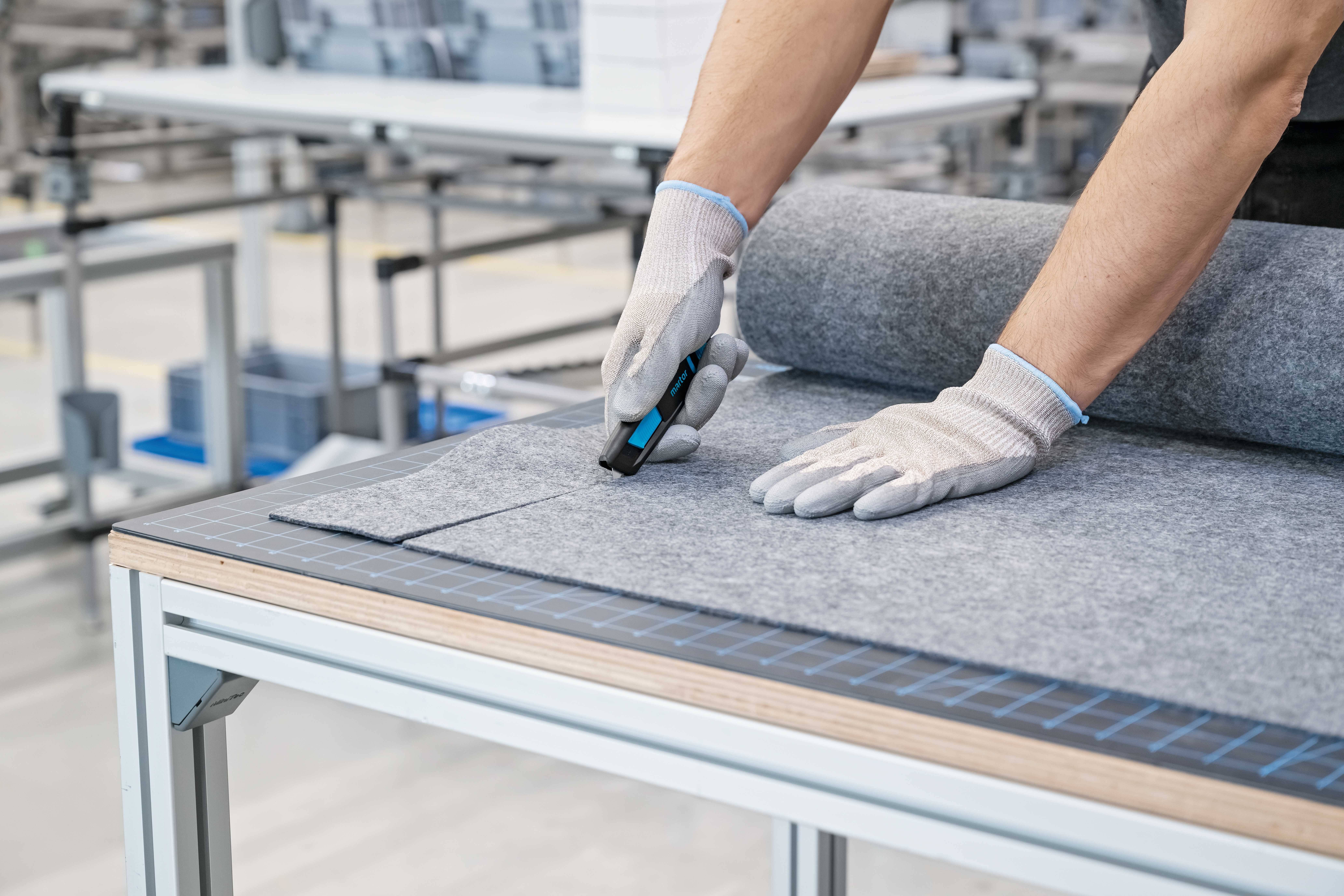 Eine Person mit Handschuhen schneidet mit einem Schneidwerkzeug ein großes graues Wollmaterial auf einem Tisch ab.