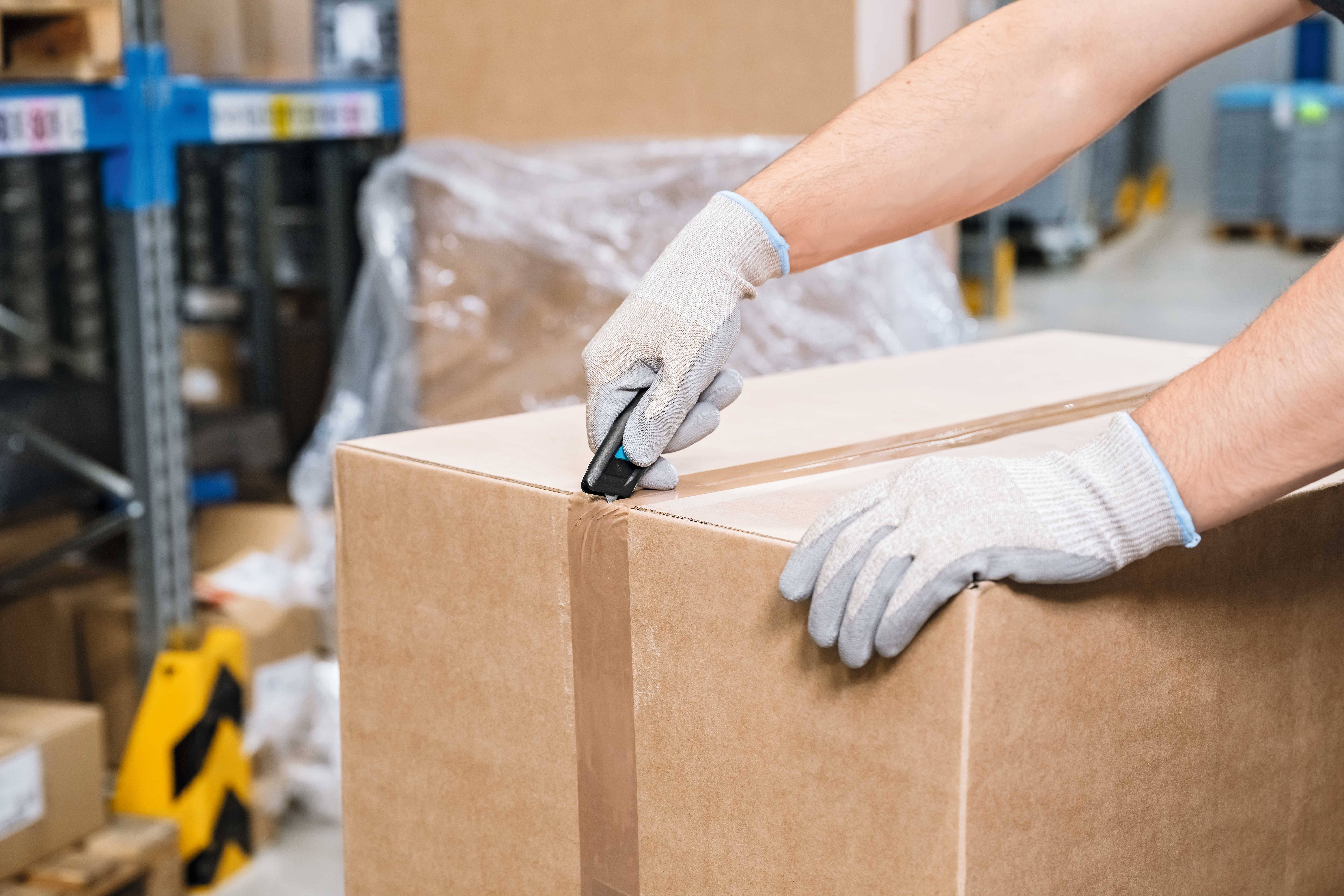 Eine Person mit Handschuhen öffnet mit einem Cutter ein großes, mit Klebeband gesichertes Paket in einem Lager.