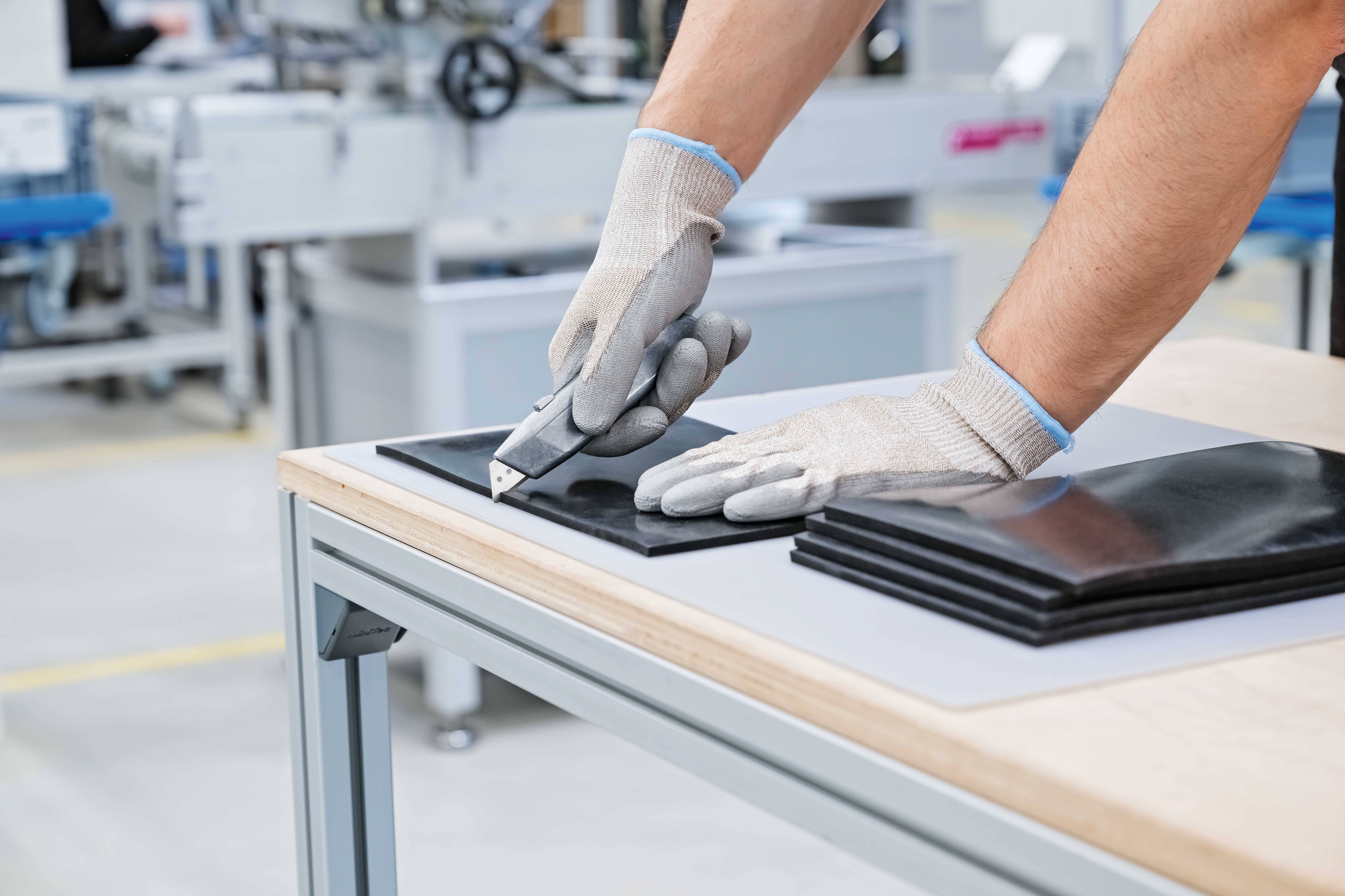 Eine Person schneidet mit einem Teppichmesser schwarze Gummiblätter auf einem Holztisch. Sie trägt graue Handschuhe.