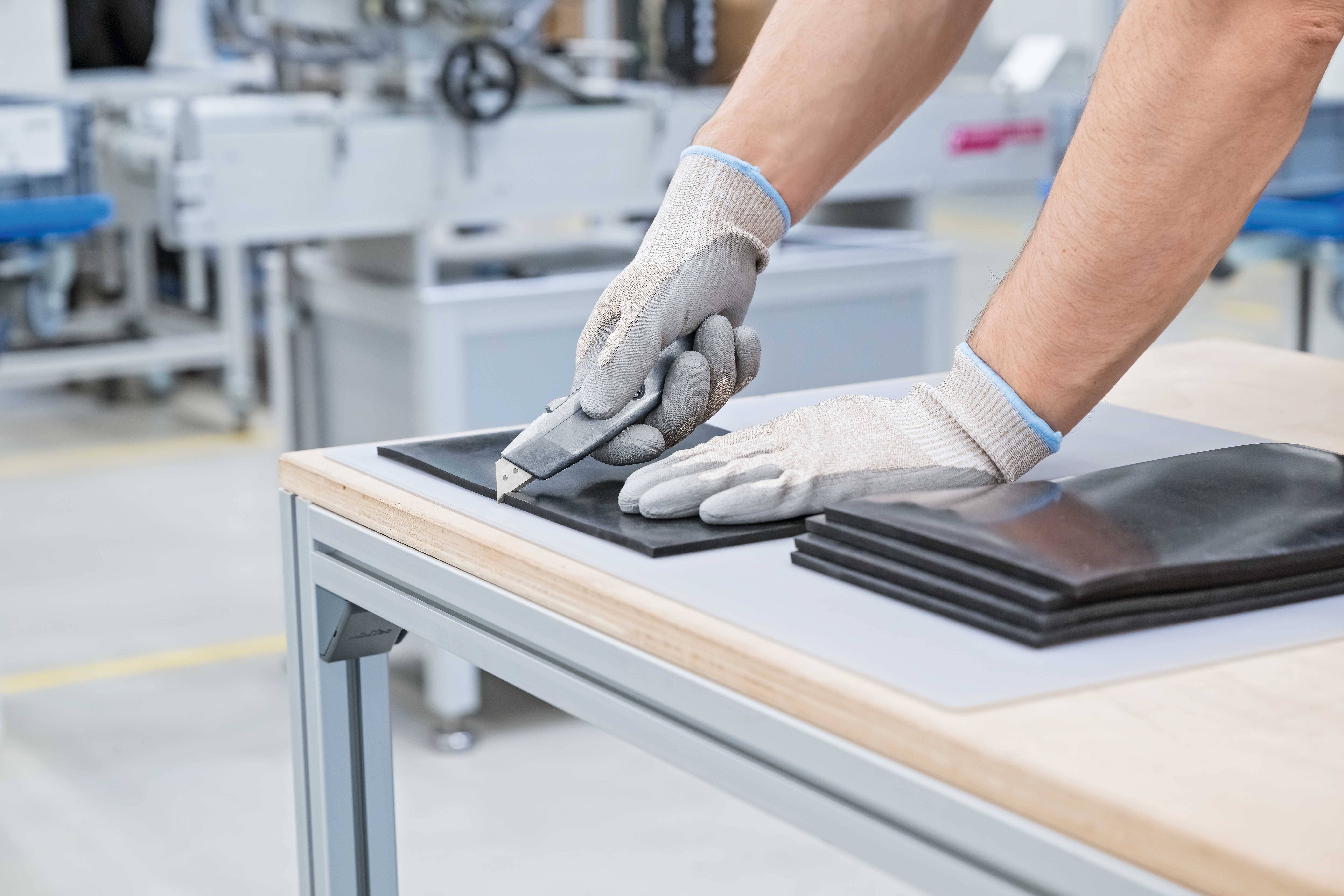 Ein Arbeiter mit Handschuhen schneidet mit einem Messer Gummimaterial auf einem Tisch in einer Fabrikumgebung.