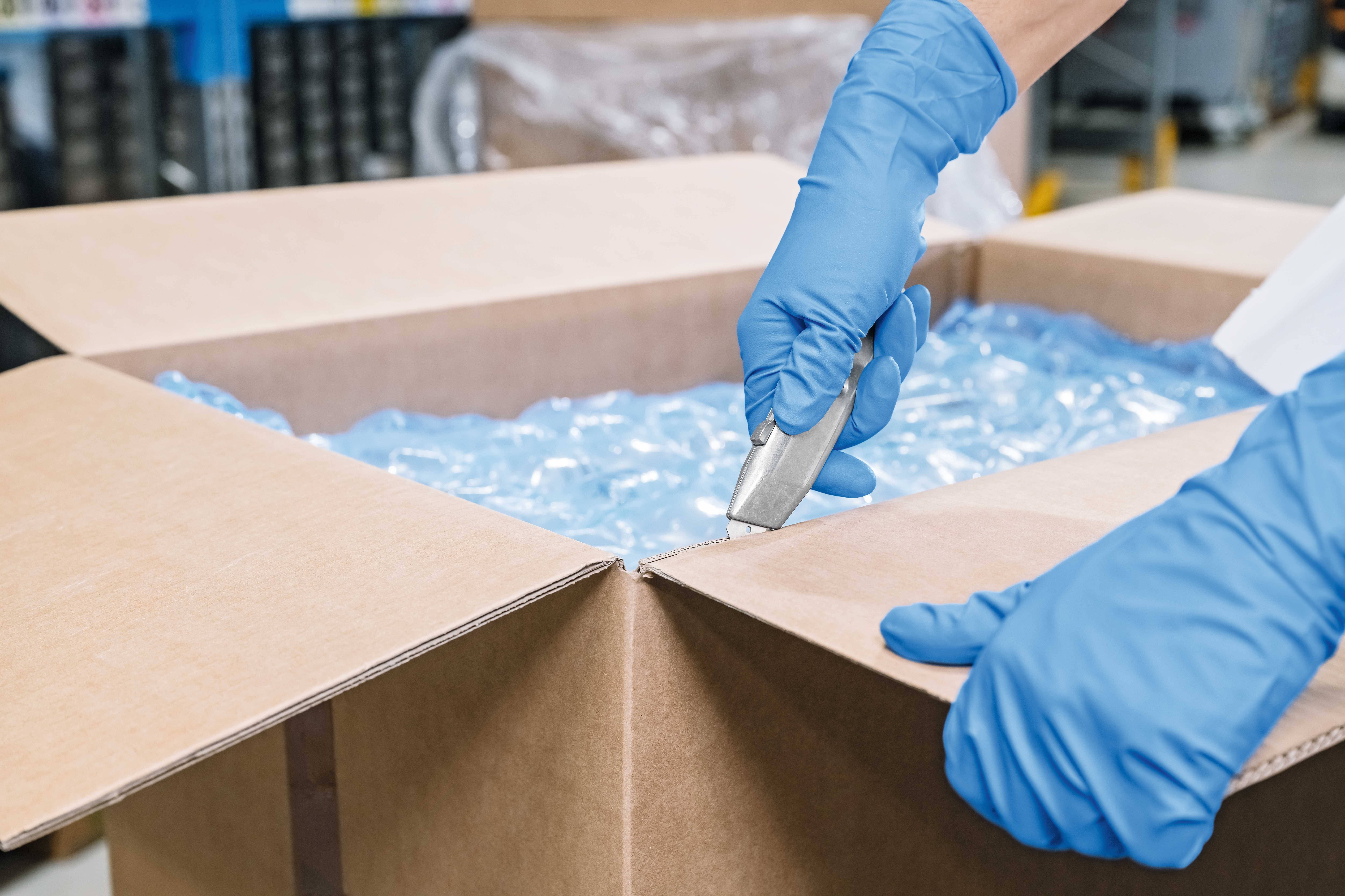 Person mit blauen Handschuhen schneidet vorsichtig einen Karton mit einem Cuttermesser auf. Verpackungsmaterial ist sichtbar.