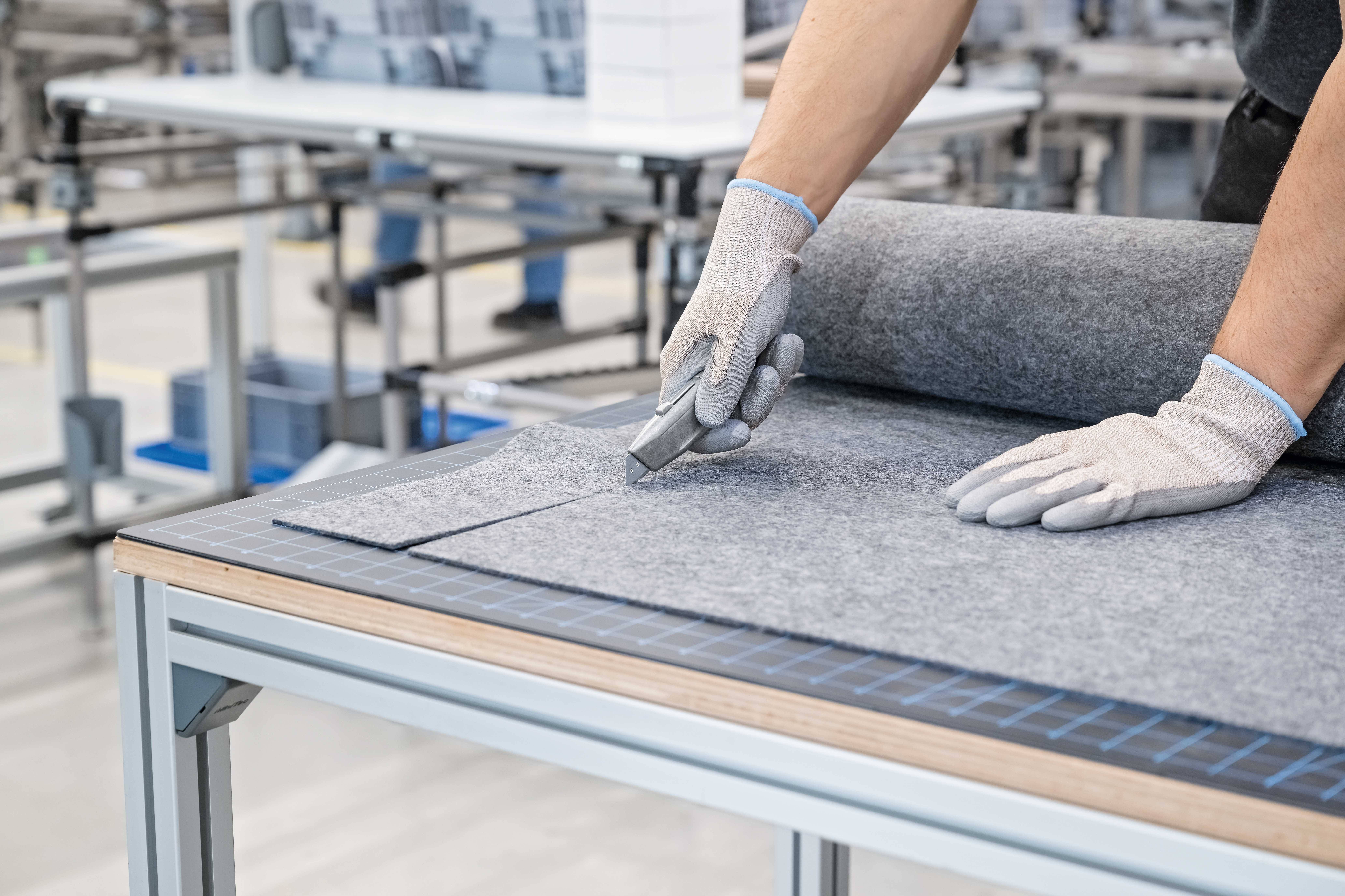 Eine Person mit Handschuhen schneidet grauen Stoff auf einem Tisch in einer Fabrikumgebung. Fokus liegt auf präzise Handarbeit.