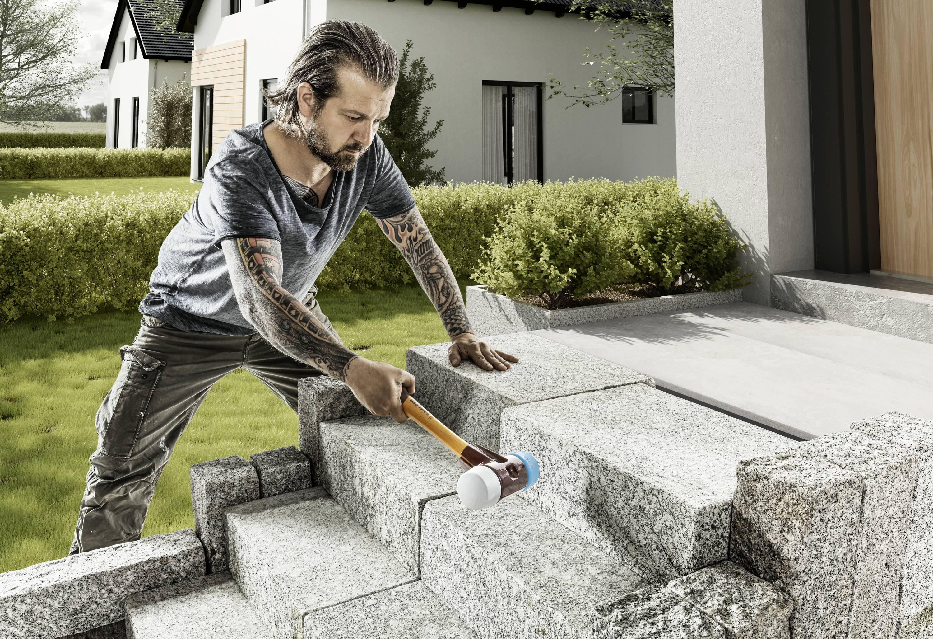 Ein Mann mit tätowierten Armen bearbeitet mit einem Hammer die Steintreppe eines modernen Hauses im Freien.