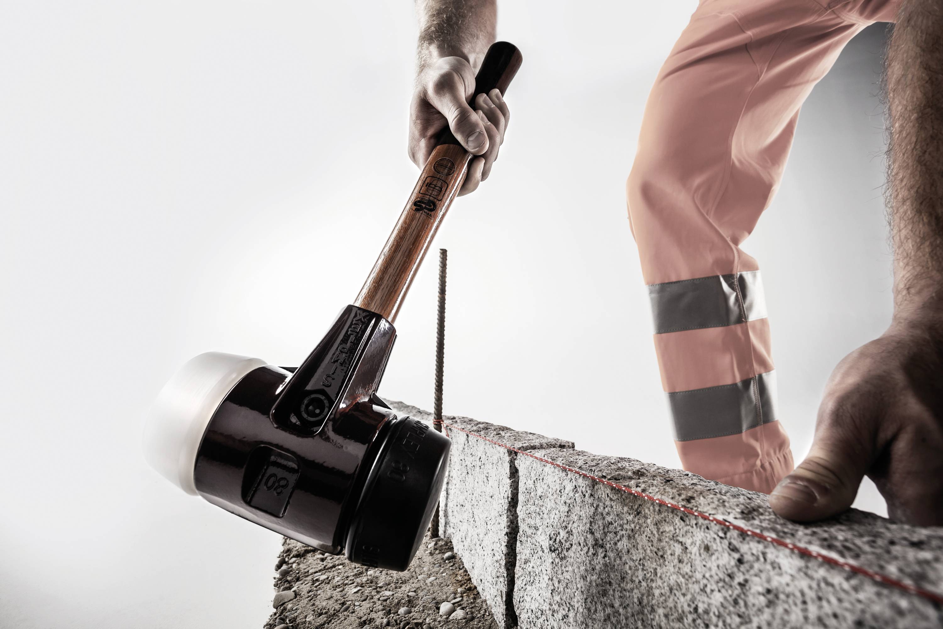 Eine Person in Arbeitskleidung schlägt mit einem Hammer auf einen Markierungspfahl auf einer Steinmauer, sorgfältig platzierend.