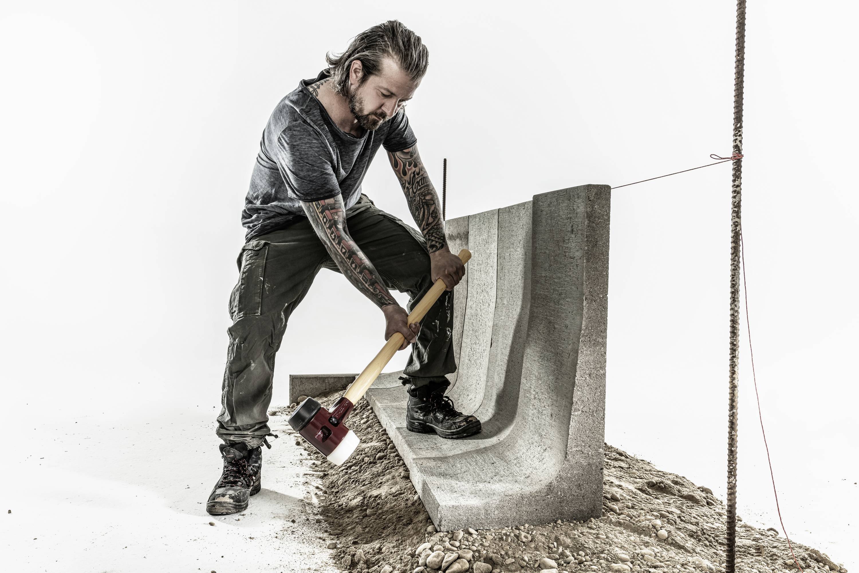 Ein Mann mit Tattoos nutzt einen großen Hammer, um einen Betonblock zu bearbeiten. Er trägt Arbeitskleidung und steht auf erdigem Boden.