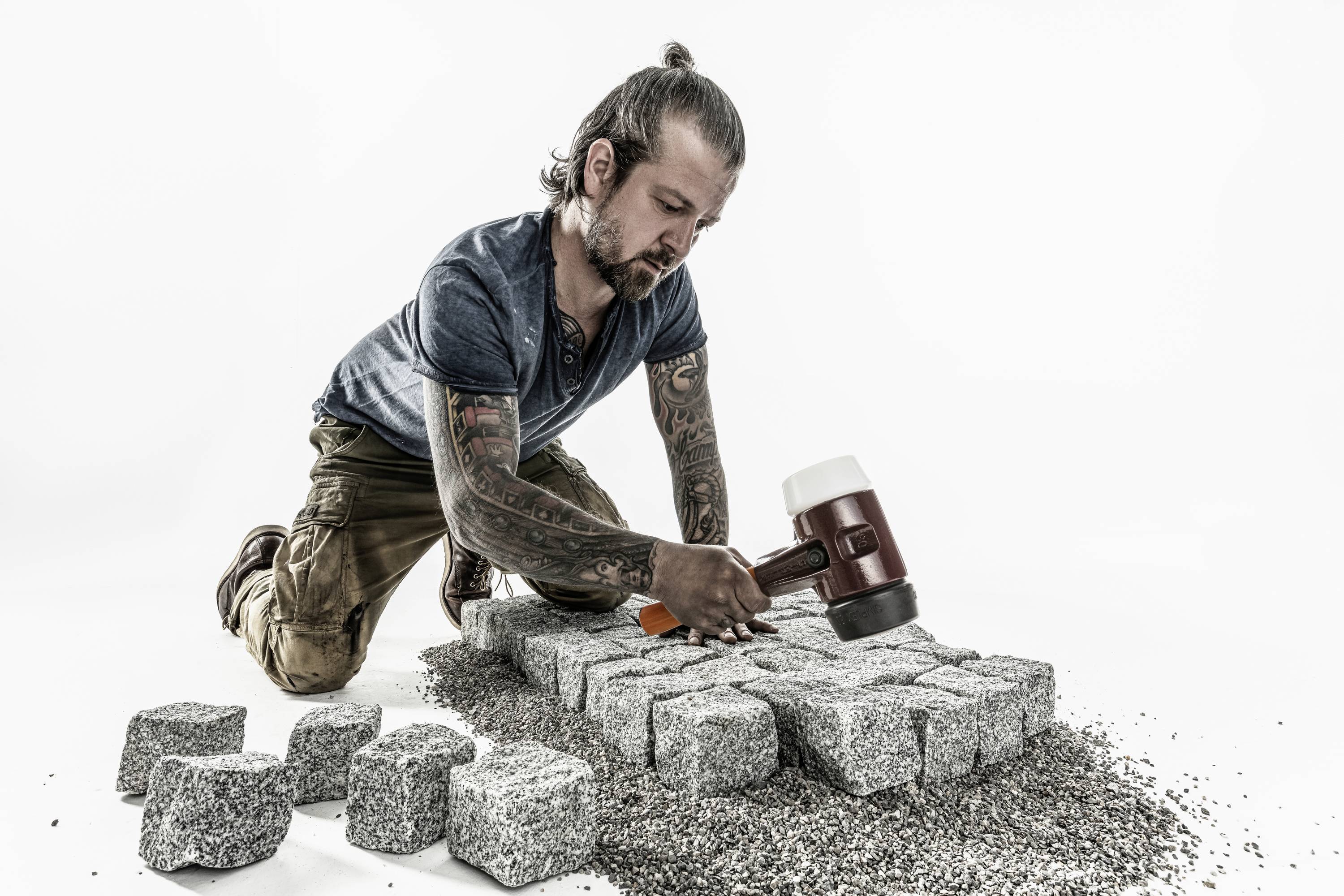 Ein Mann mit Tattoos kniet auf dem Boden und verlegt Pflastersteine. Er hält einen Gummihammer und trägt ein blaues Hemd und beige Hosen.