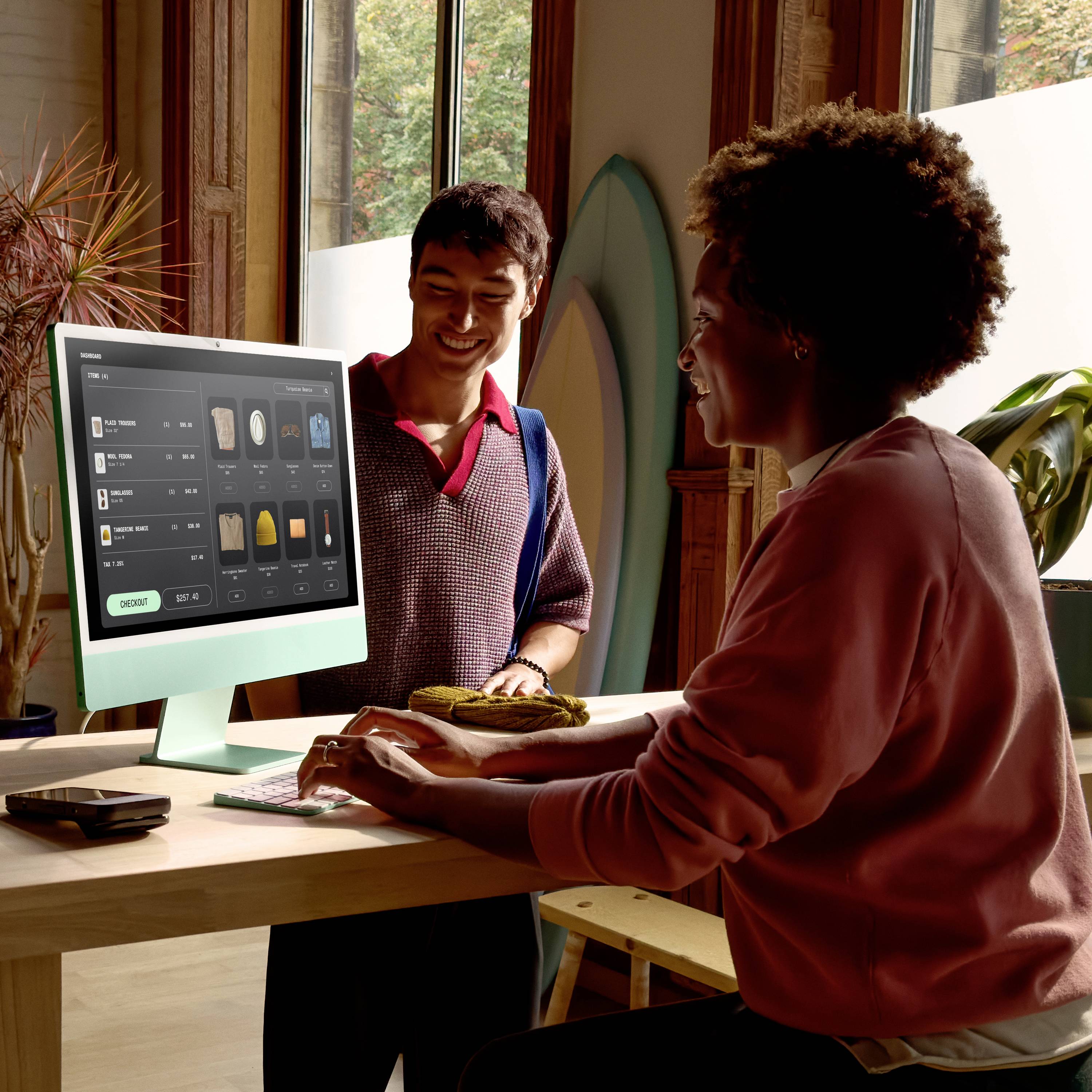 Zwei Personen in einem modernen Café bestellen online an einem Computer auf einem Holzschreibtisch, mit Pflanzen im Hintergrund.