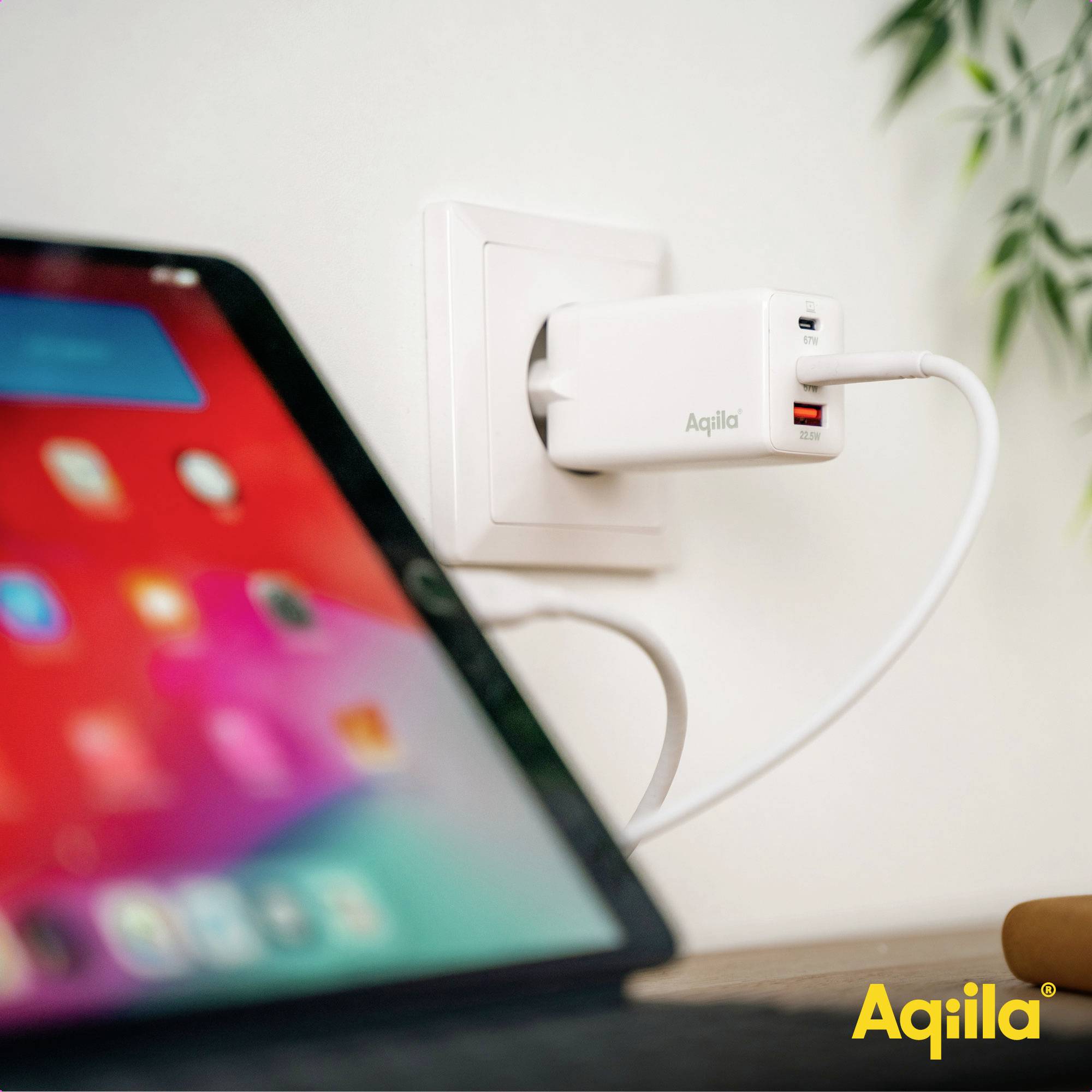 Ein Handy lädt mithilfe eines Aquila-Ladegeräts, das in einer Steckdose steckt. Ein Tablet ist in der Nähe sichtbar.