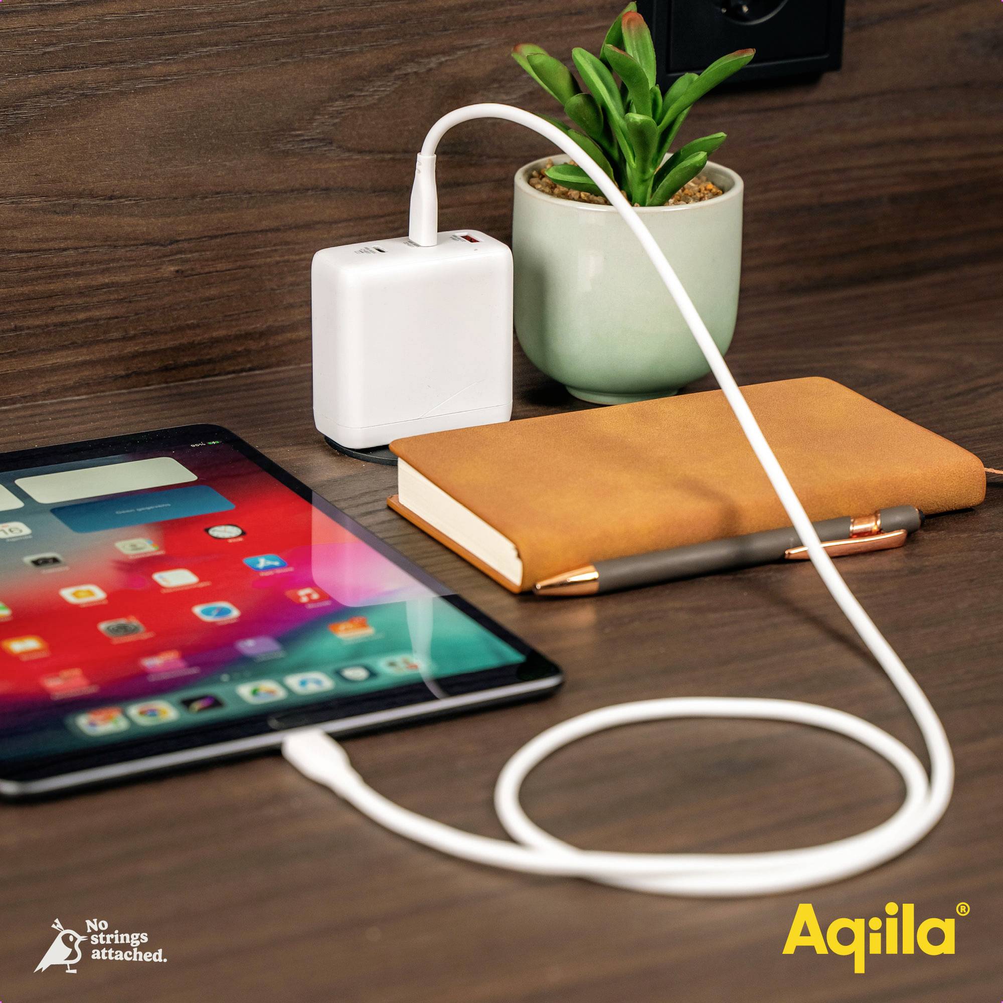 Ein Tablet wird über ein weißes Ladekabel mit einem Ladegerät verbunden. Daneben liegen ein Notizbuch mit Stift und eine kleine Pflanze.