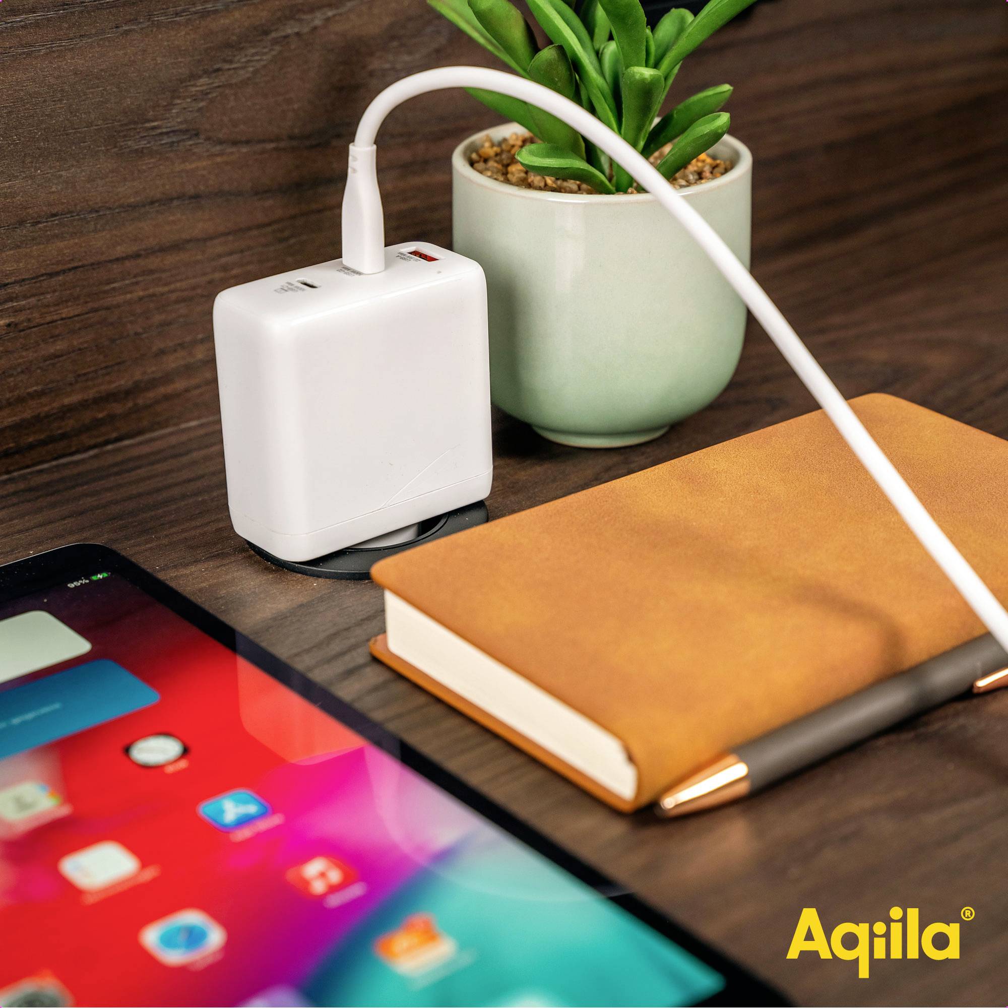 Ein Ladegerät und ein Notizbuch auf einem Tisch neben einem Tablet. Eine grüne Pflanze steht daneben. Branding-Logo 'Aqiila' in der Ecke.