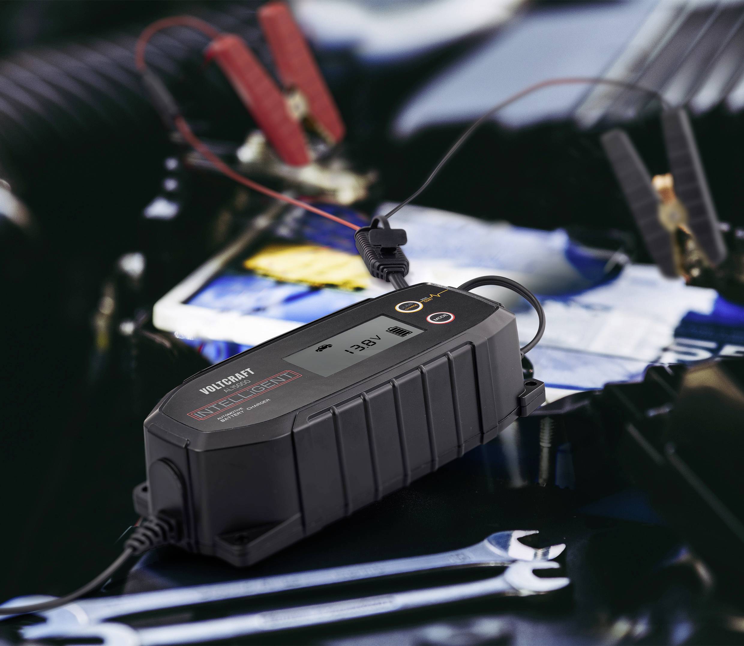 Ein Batterieladegerät ist mit Autobatterieklemmen verbunden. Es zeigt eine Spannung von 13,6V an. Im Hintergrund ist ein unscharfer Motorraum zu sehen.