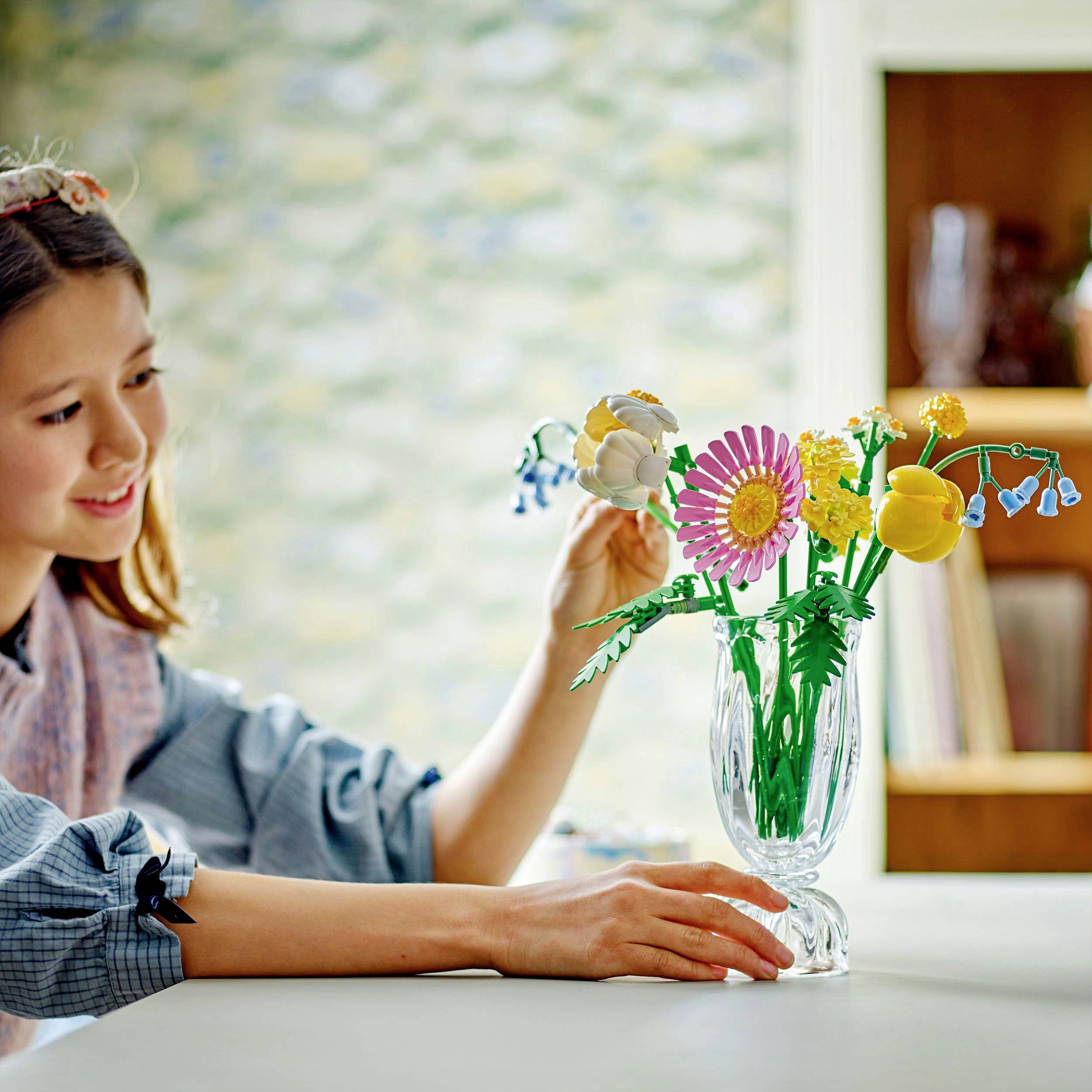 Ein Mädchen arrangiert leuchtend bunte Origami-Blumen in einer Vase auf einem Tisch. Im Hintergrund ist ein Bücherregal zu sehen.