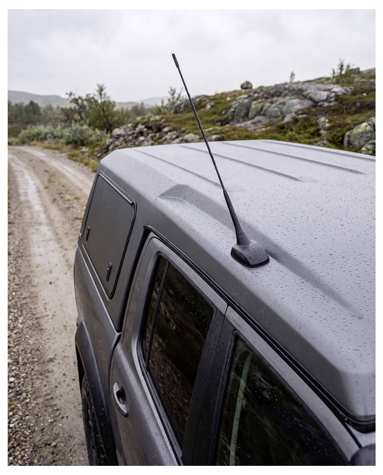 Oben auf einem grauen Geländewagen auf einer nassen Kiesstraße, mit einer Antenne und bewölkter Landschaft im Hintergrund.