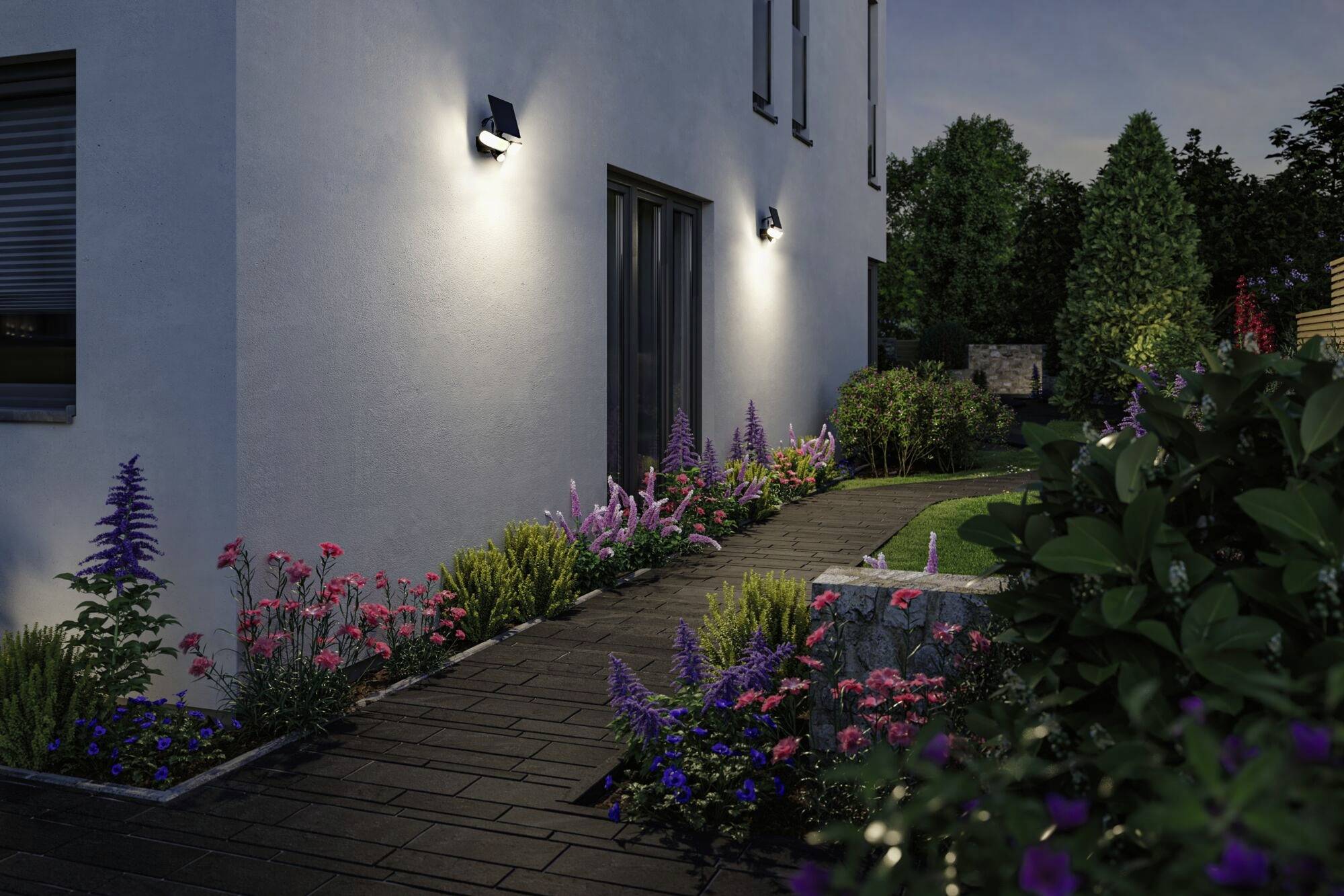 Gartenweg bei Nacht, beleuchtet durch Wandlichter; umgeben von bunten Blumen und Sträuchern entlang eines modernen Hauses.
