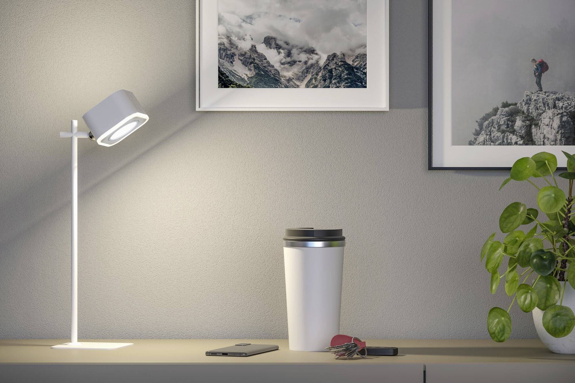 Ein Schreibtisch mit Lampe, Kaffeebecher, Smartphone und Schlüsseln darauf. An der Wand hängen Bilder, eine Pflanze rechts.