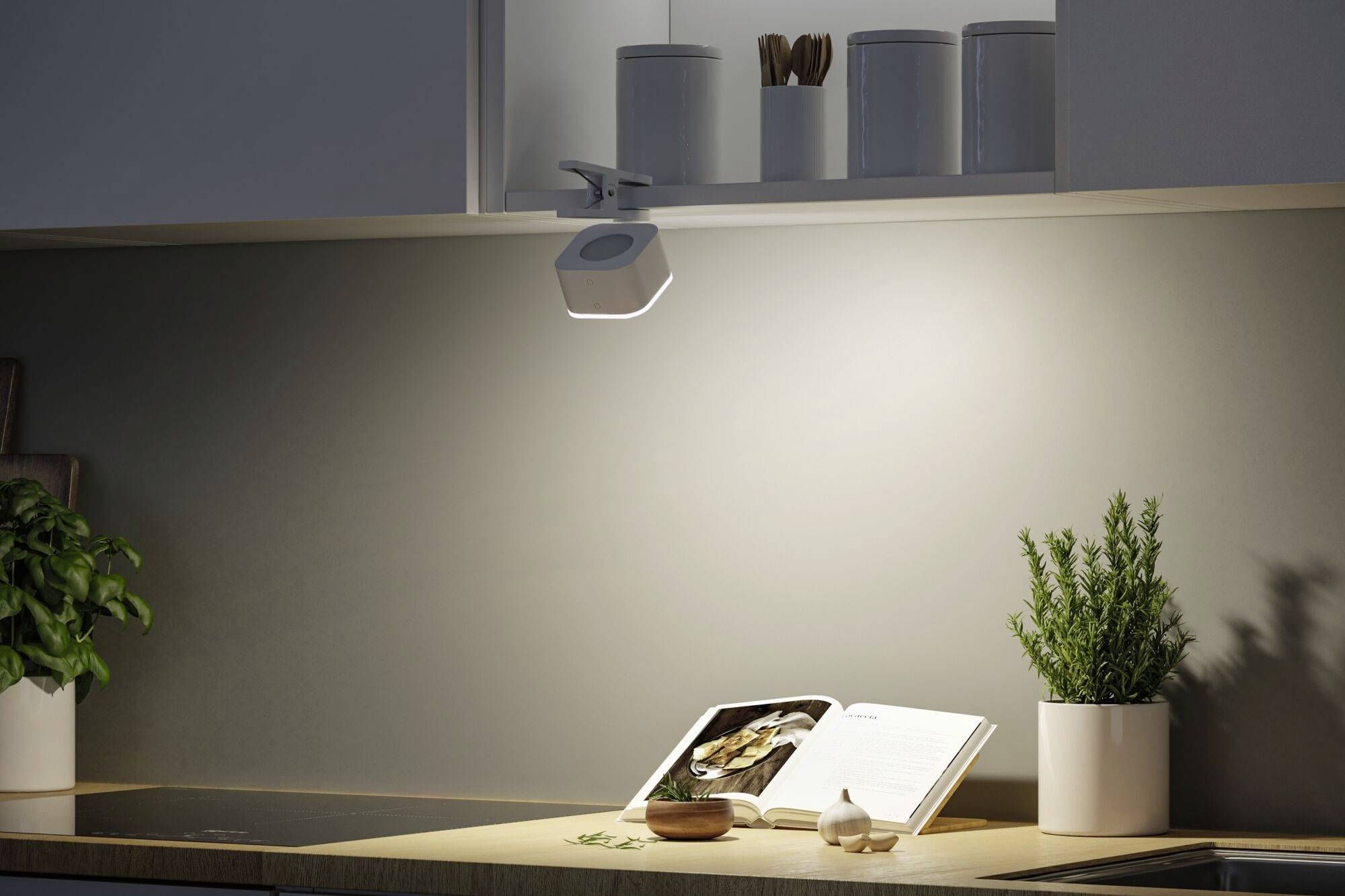 Eine moderne Küchenecke mit Lichtstrahler auf eine Arbeitsplatte, auf der ein offenes Kochbuch, ein Topf mit Kräutern und eine Knoblauchzehe stehen.