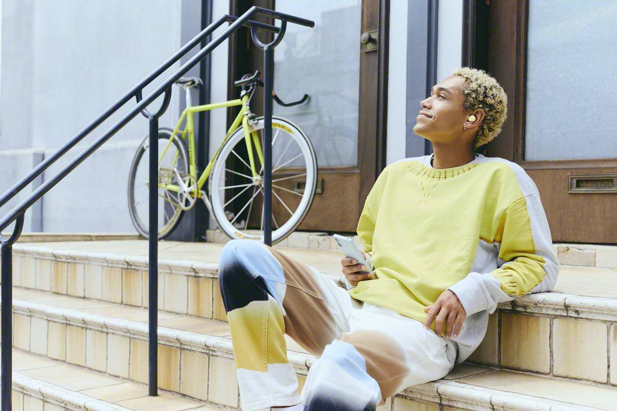 Eine Person sitzt auf einer Treppe, hört Musik über Ohrhörer und hält ein Smartphone in der Hand. Ein Fahrrad steht im Hintergrund.