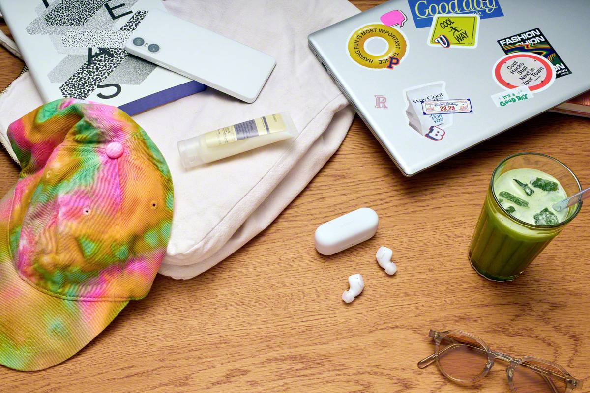 Eine bunte Kappe, Sonnenbrille, Kopfhörer und ein Matcha-Getränk liegen auf einem Tisch neben einem Laptop mit Aufklebern.