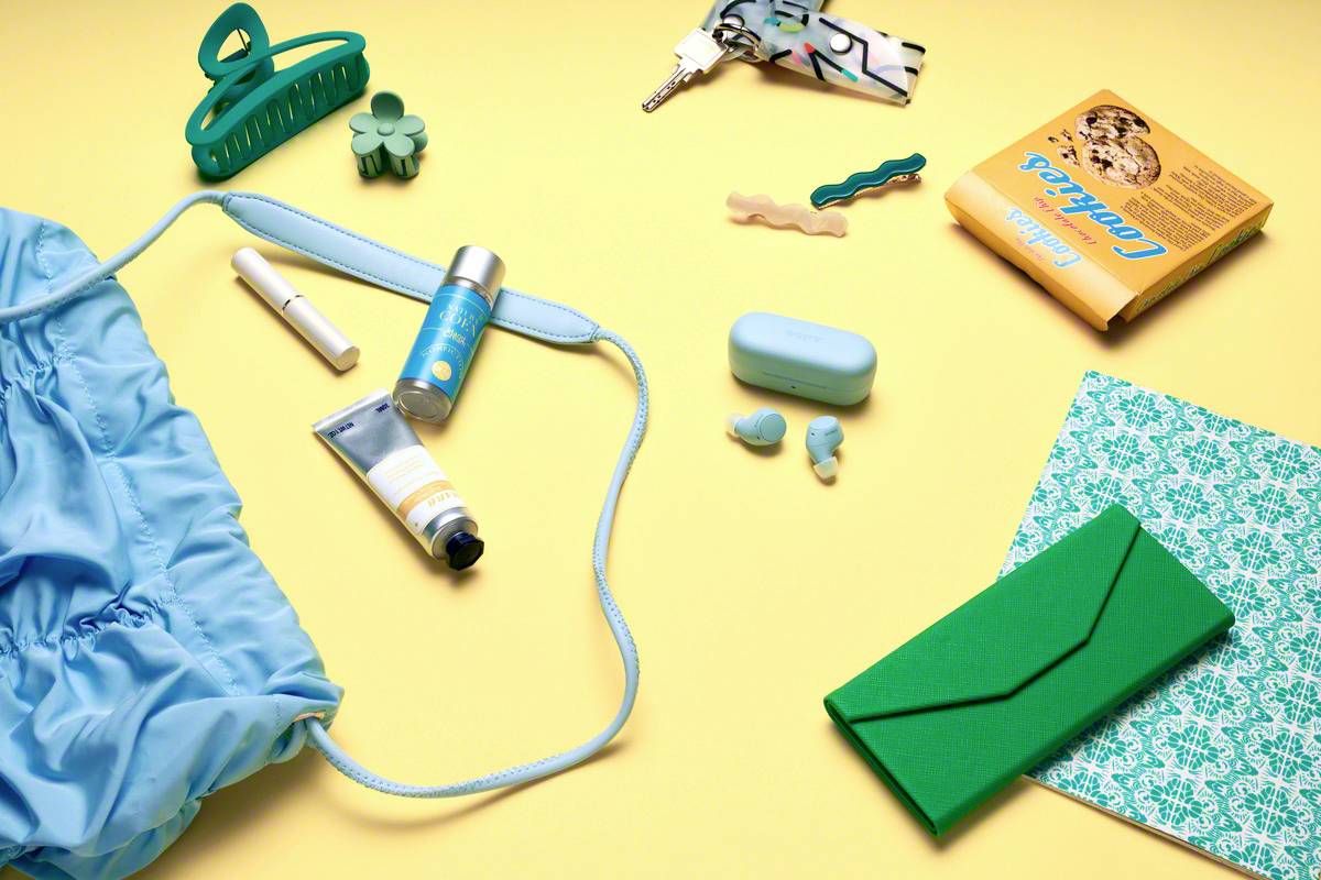 Eine blaue Tasche auf einem gelben Hintergrund, umgeben von Alltagsgegenständen wie Schlüssel, Kosmetik, einem Buch und einem grünen Notizbuch.