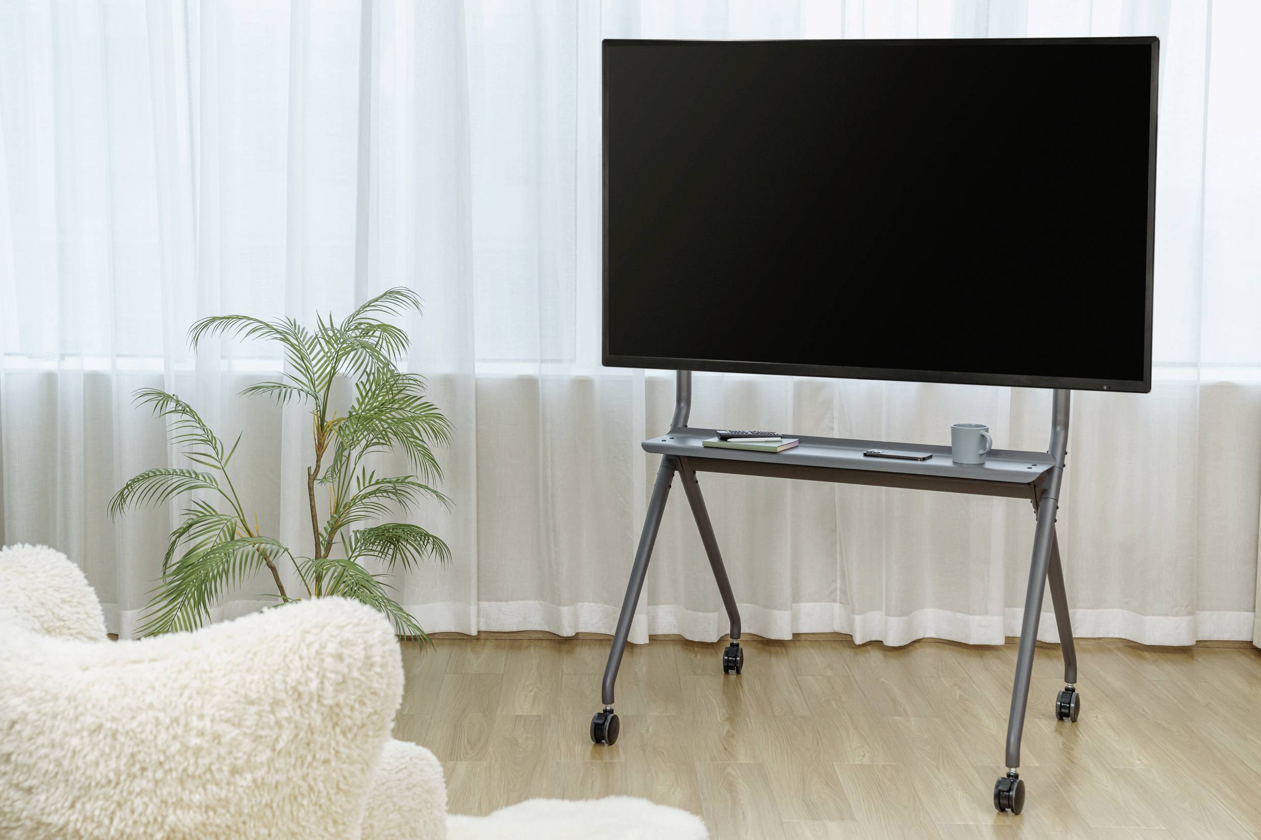 Flachbildfernseher auf rollbarem Ständer in einem hellen Raum mit Holzboden. Ein Sessel und eine Pflanze sind im Hintergrund.