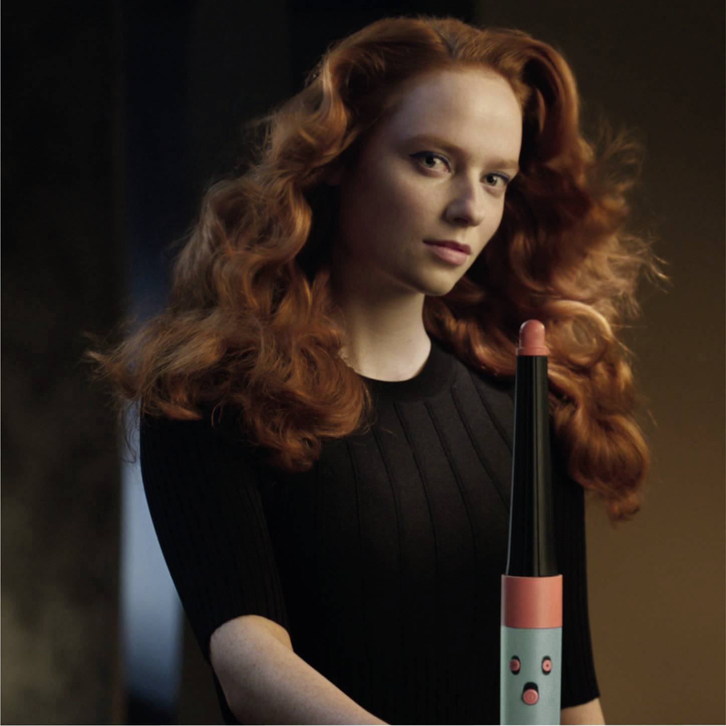 Eine Frau mit roten Haaren steht vor einem neutralen Hintergrund, hält einen modernen, länglichen Gegenstand.