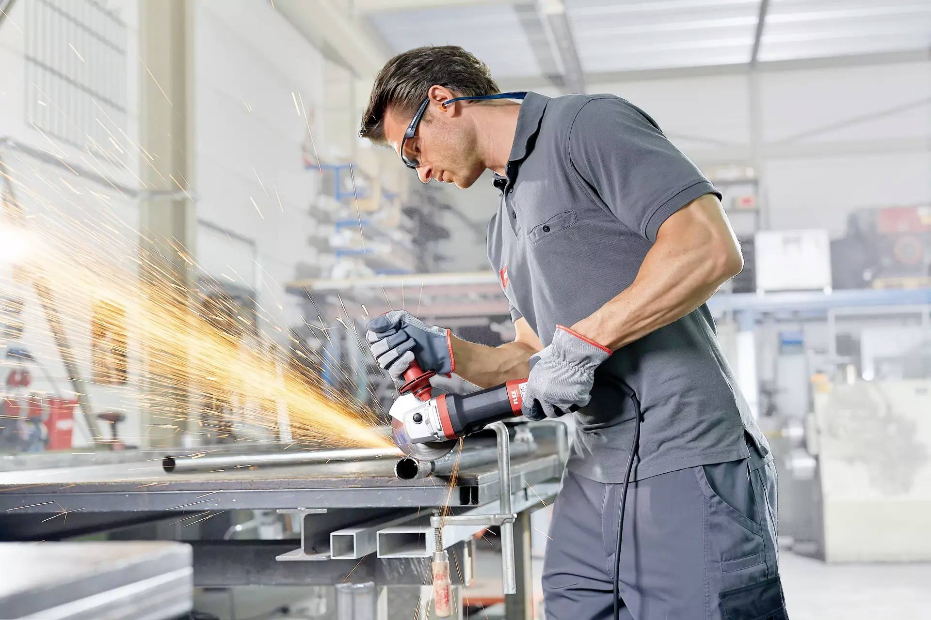 Ein Mann trägt Schutzbrille und schleift in einer Werkstatt ein Metallstück mit einem Winkelschleifer, wodurch Funken entstehen.