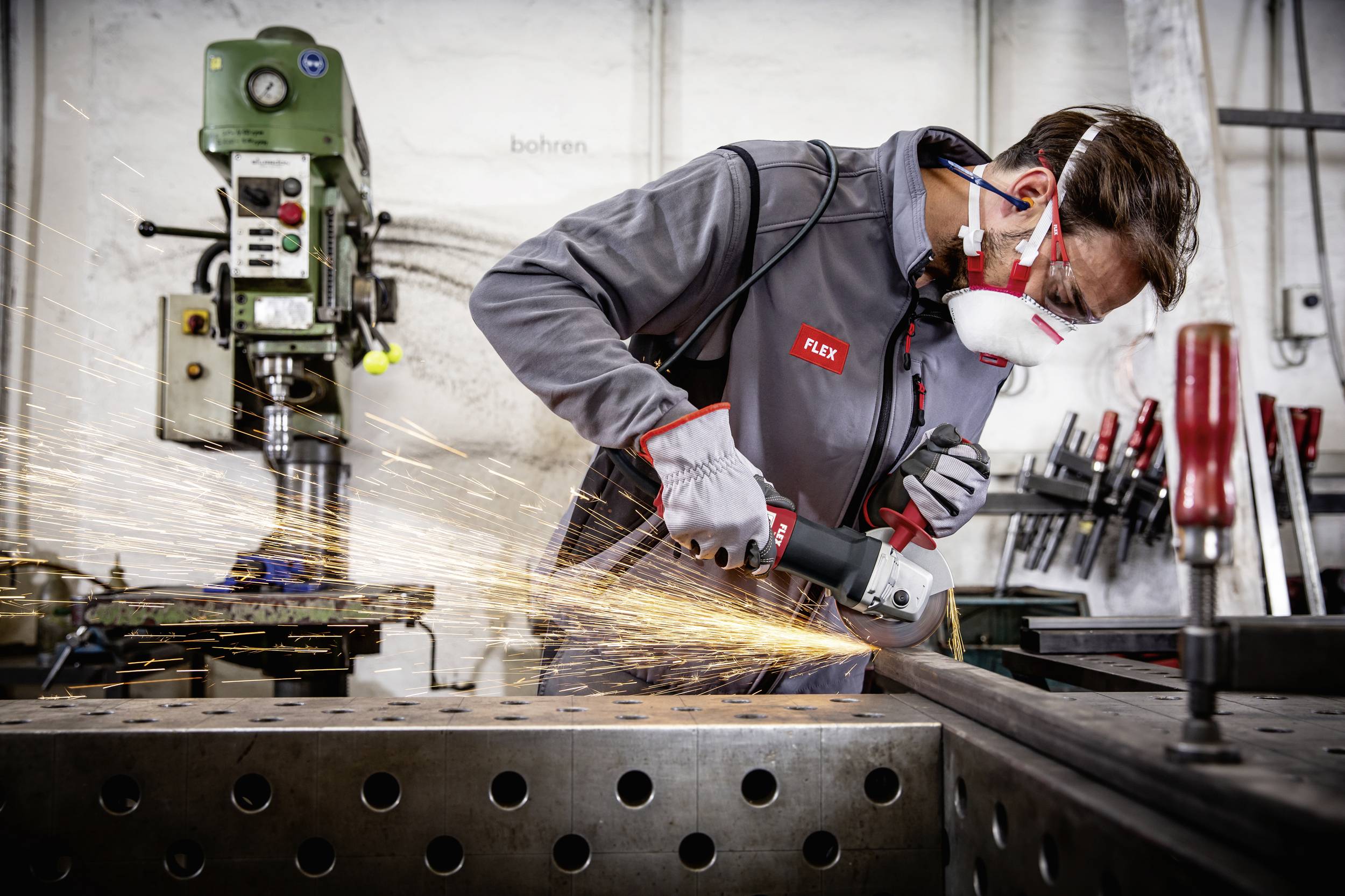 Ein Mann arbeitet mit einem Winkelschleifer an einer Metallplatte. Funken fliegen. Er trägt Schutzkleidung und Atemschutz.