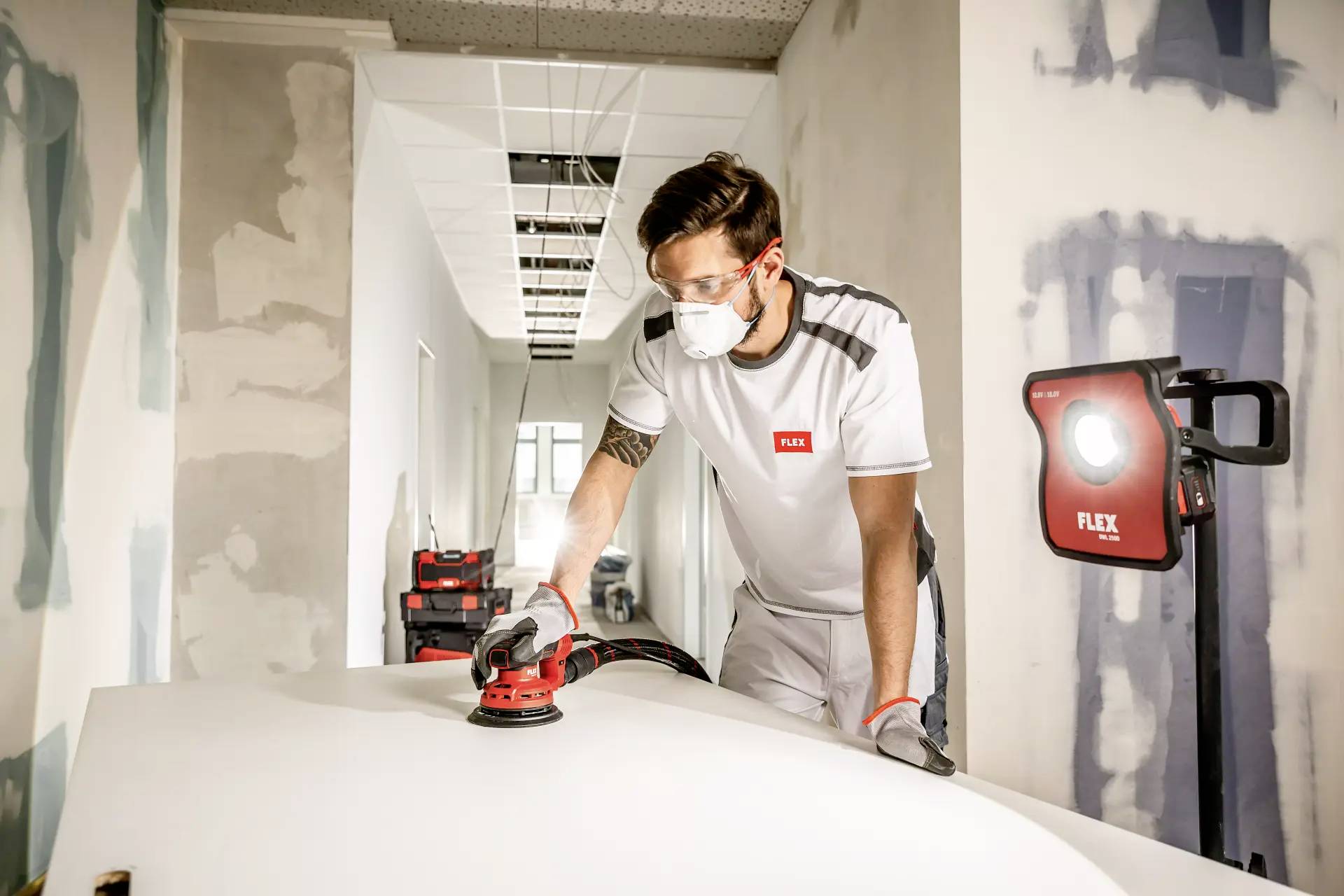 Ein Mann in Arbeitskleidung schleift eine weiße Oberfläche mit einem Elektrowerkzeug in einem halb renovierten Raum. Er trägt Schutzbrille und Maske.