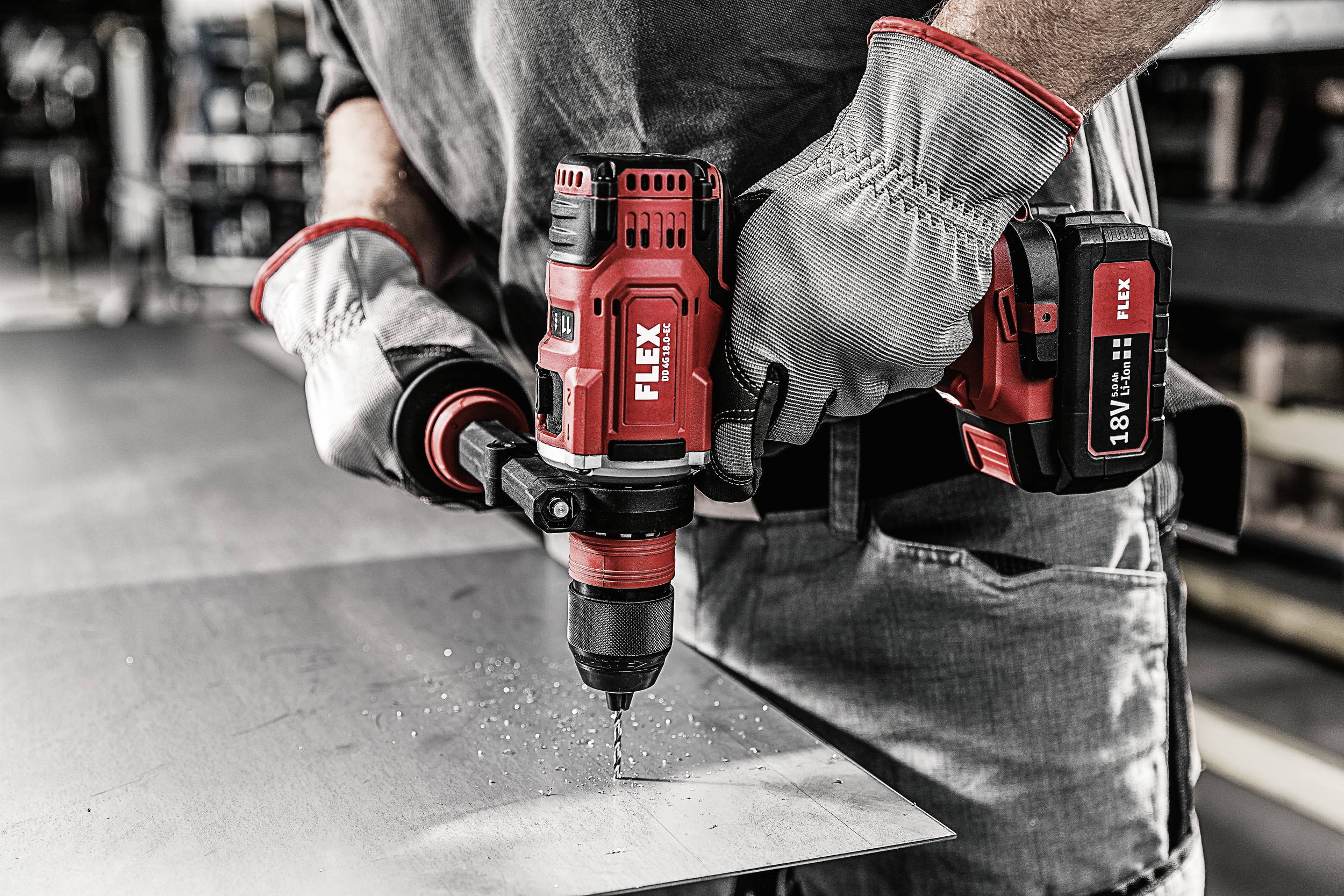 Person, die mit einem roten Akku-Bohrschrauber in eine Metallplatte bohrt, Hände in Schutzhandschuhen. Konzentration auf das Werkzeug.