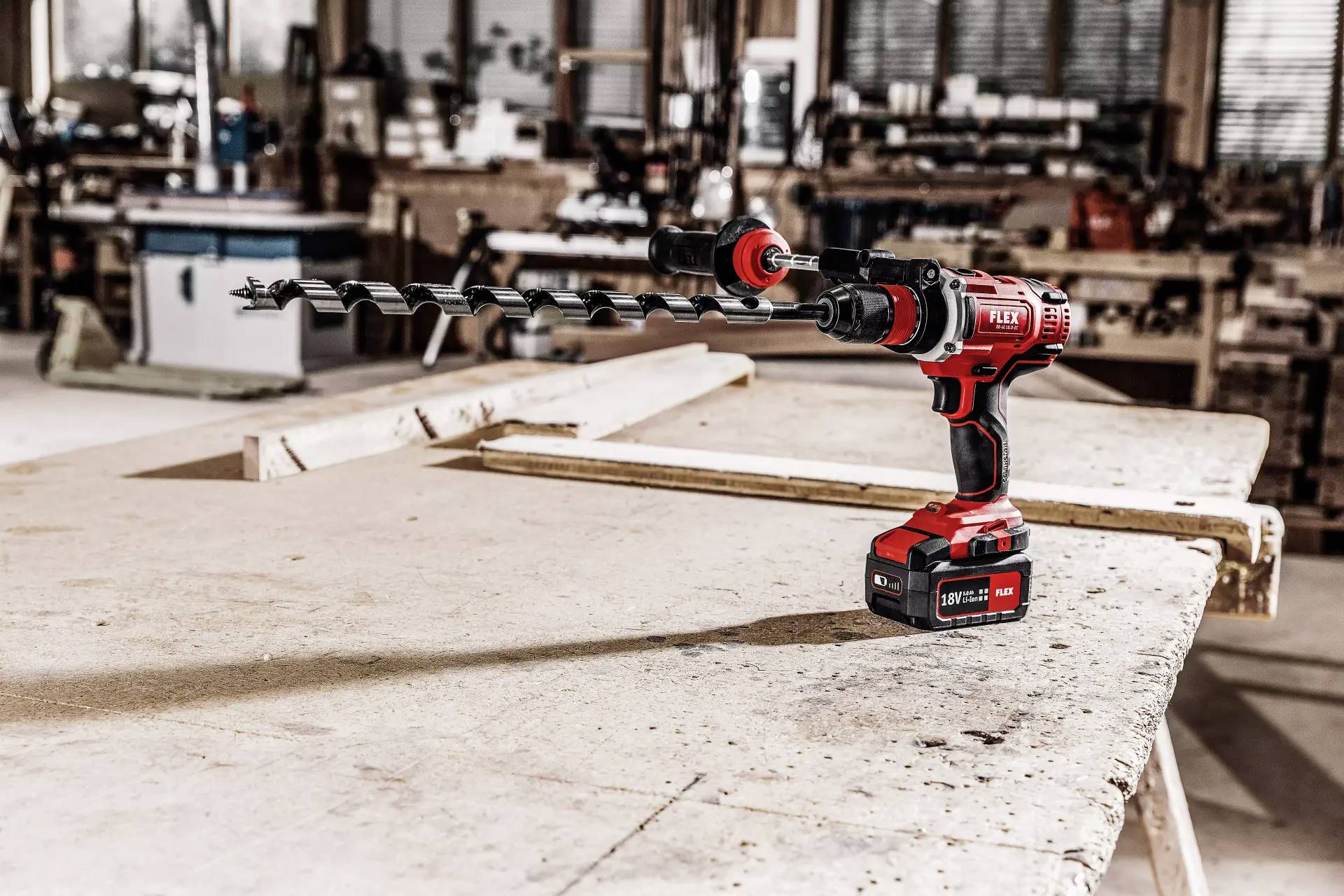 Akkubohrmaschine mit langem Bohrer auf Werkbank in einer Werkstatt; im Hintergrund unscharfe Werkstattgeräte.