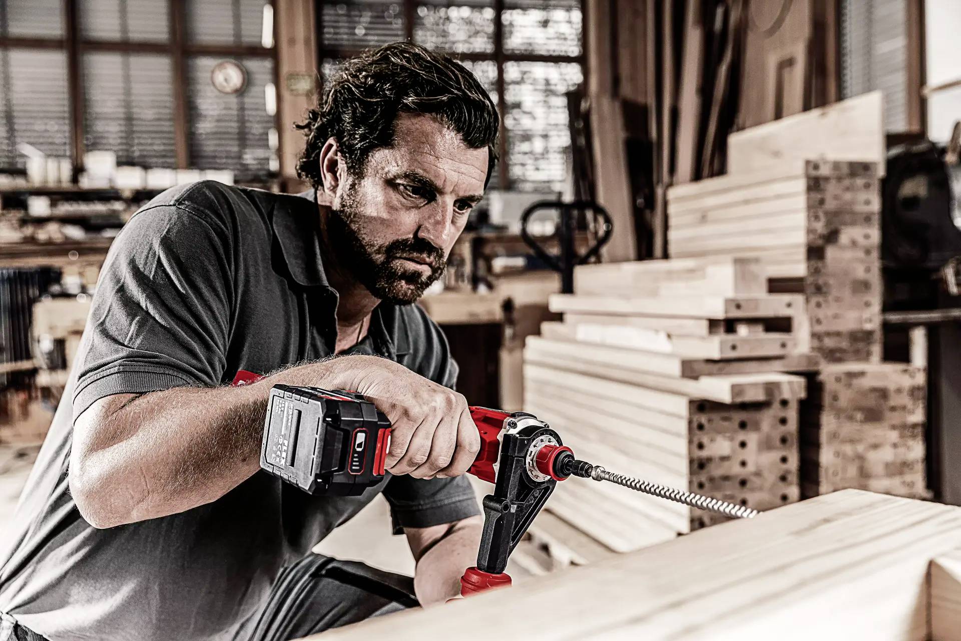 Ein Mann in einer Werkstatt nutzt eine elektrische Bohrmaschine, um Holz zu bearbeiten. Im Hintergrund sind Holzstapel zu sehen.