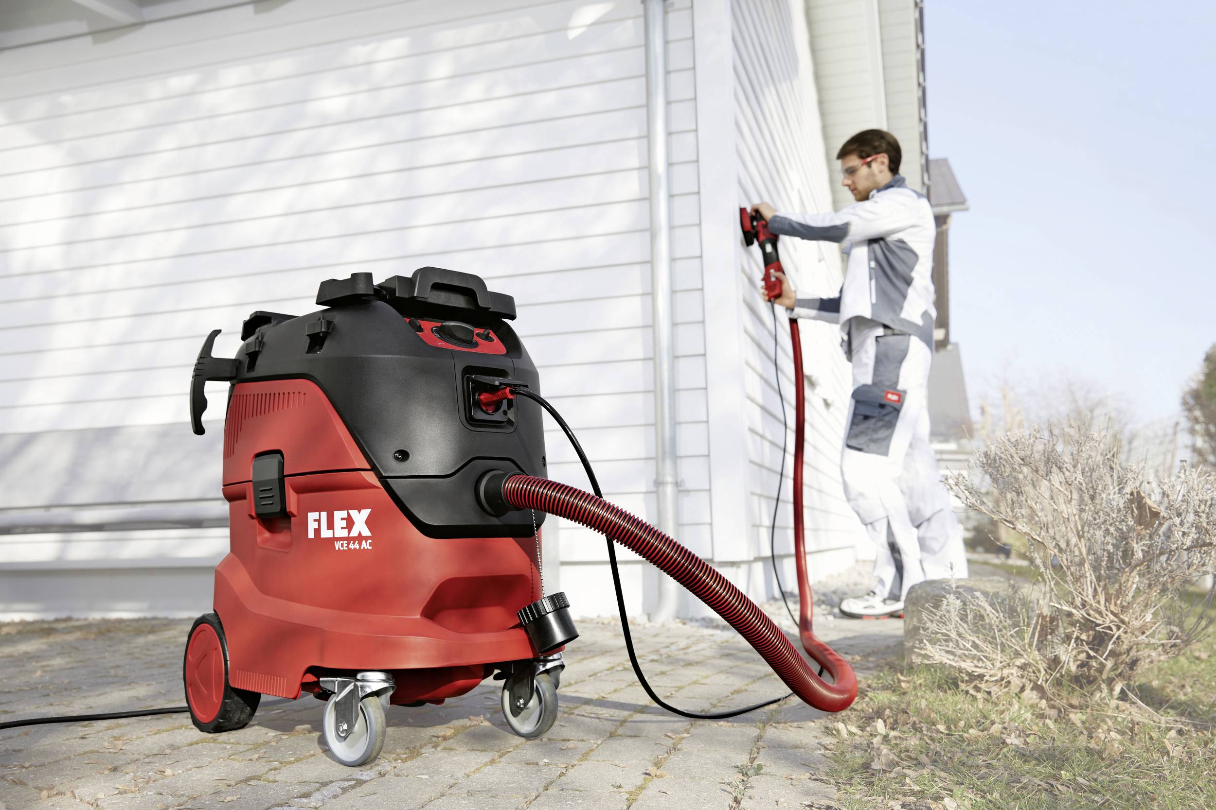 Ein roter Industriesauger der Marke Flex steht im Vordergrund. Im Hintergrund bearbeitet eine Person die Außenwand eines Gebäudes.
